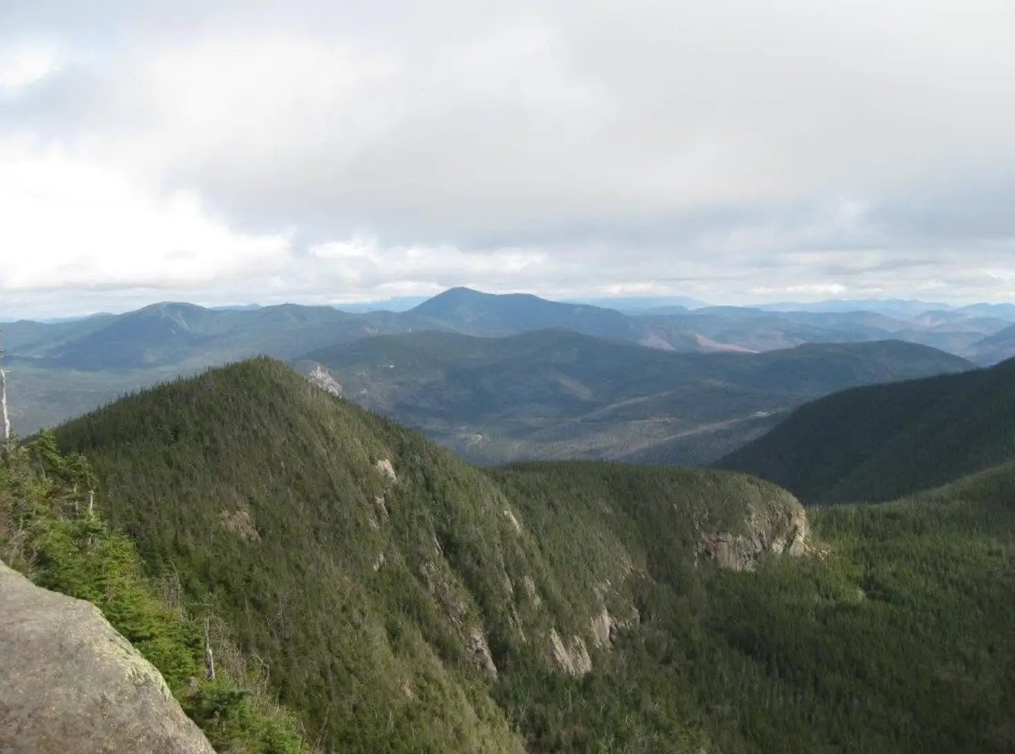 Forested mountain ridges stretch into the distance beneath a cloudy sky from a rocky overlook.