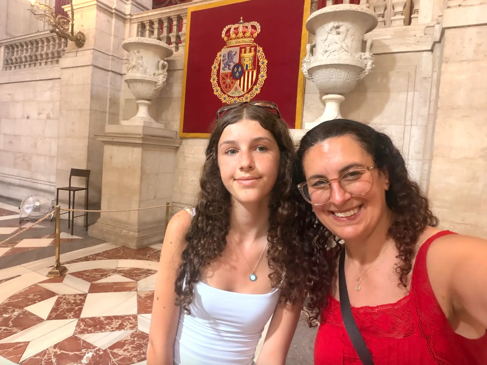 Two visitors pose for a selfie inside an ornate marble hall beneath a red banner with the Spanish coat of arms.