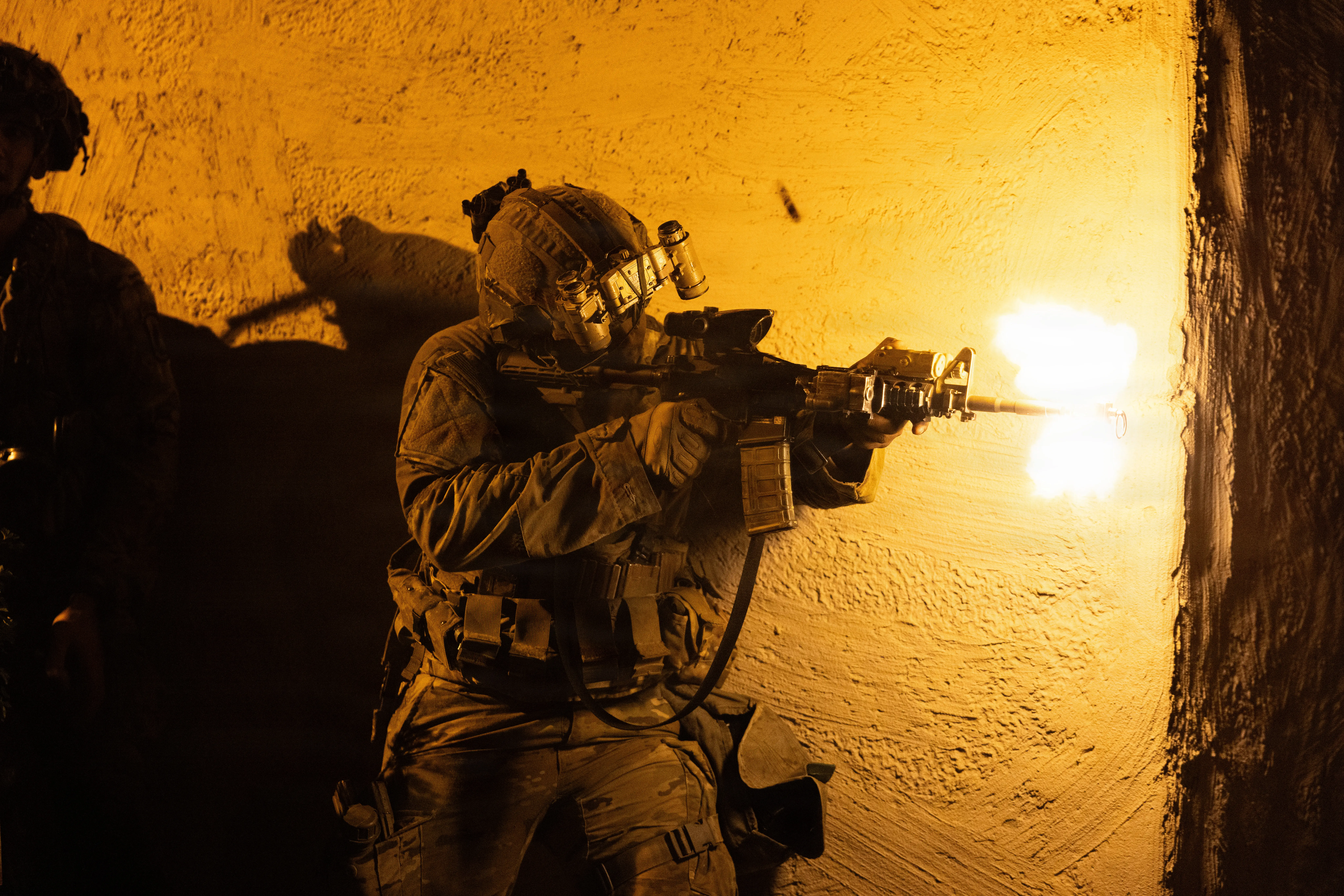 A US Army soldier fires a rifle while standing next to a white wall.