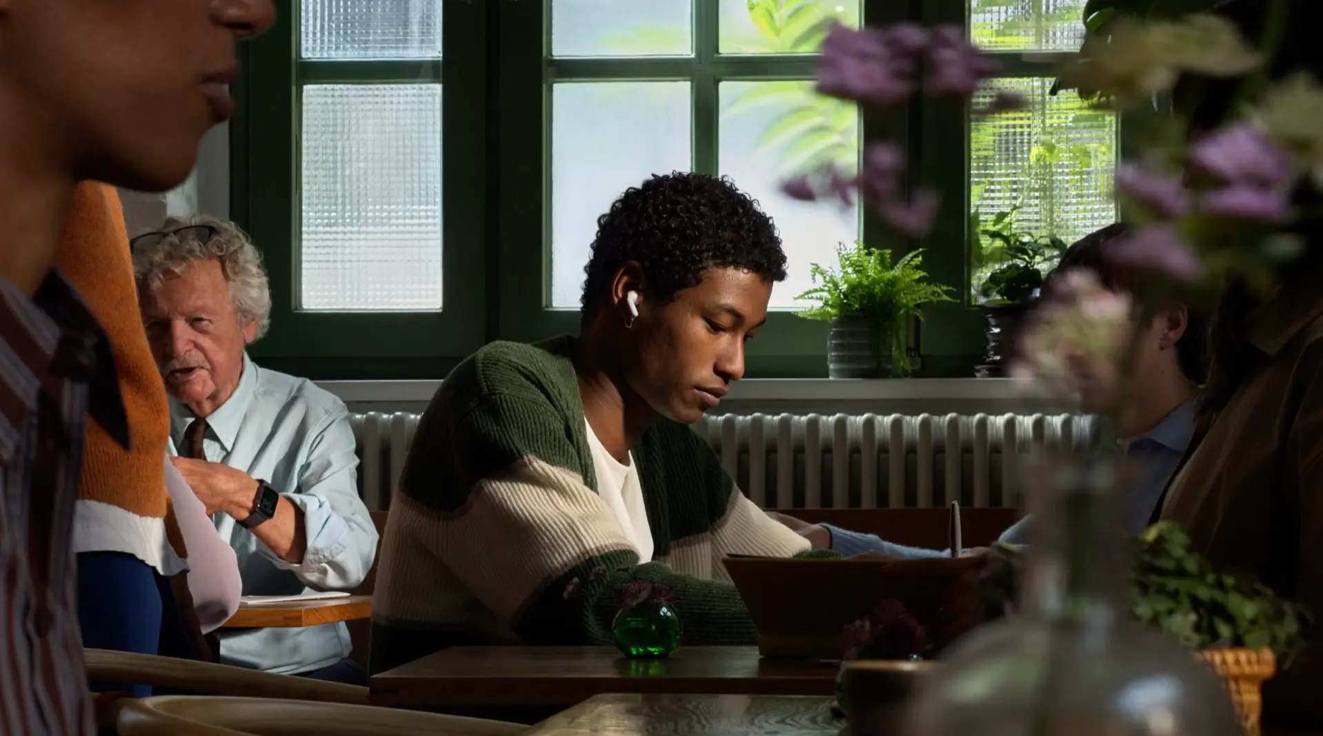 An AirPods 5 user sits at a table at a busy cafe.
