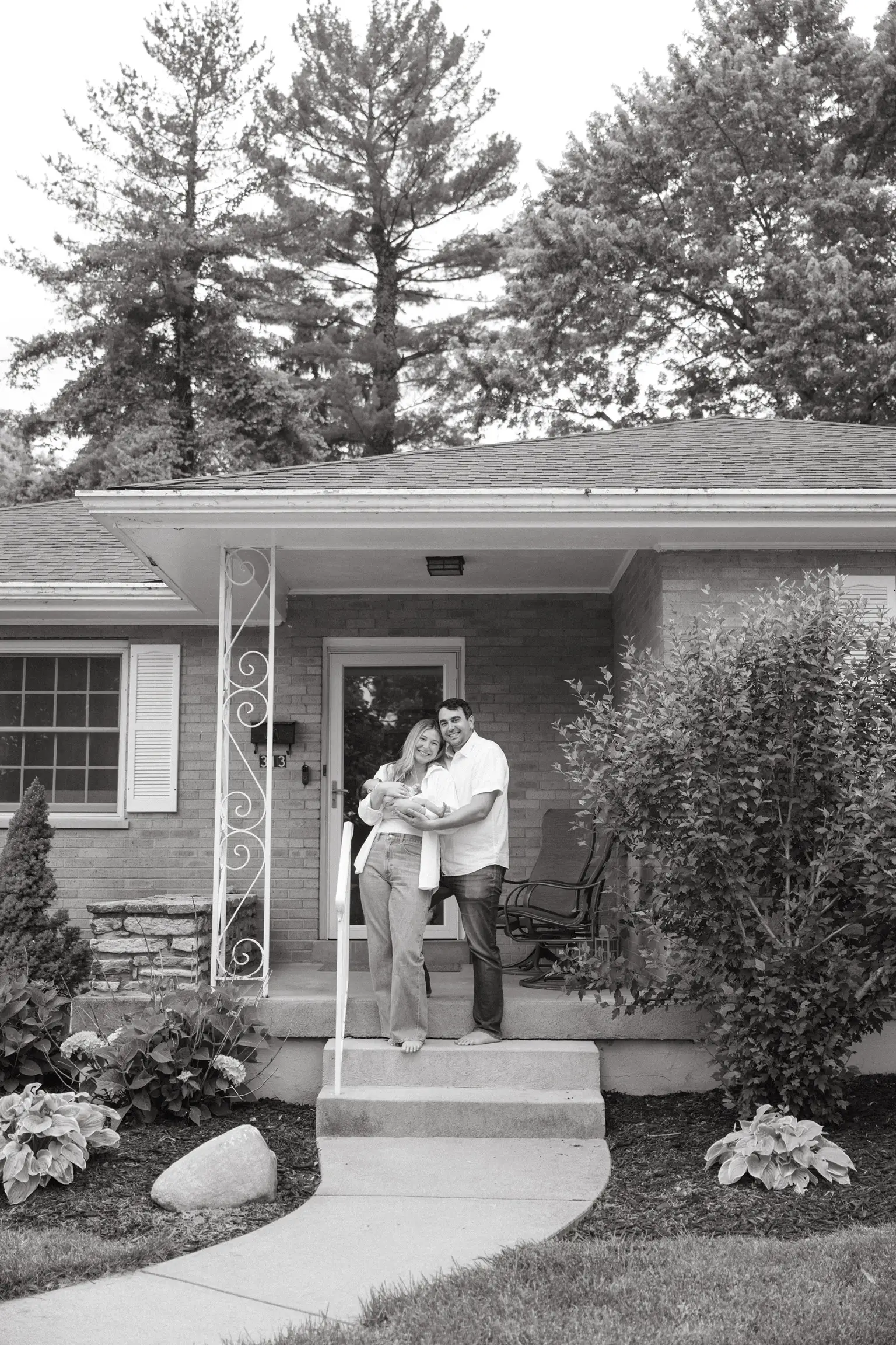 Two adults stand on a brick home's front porch holding a swaddled baby in black and white.
