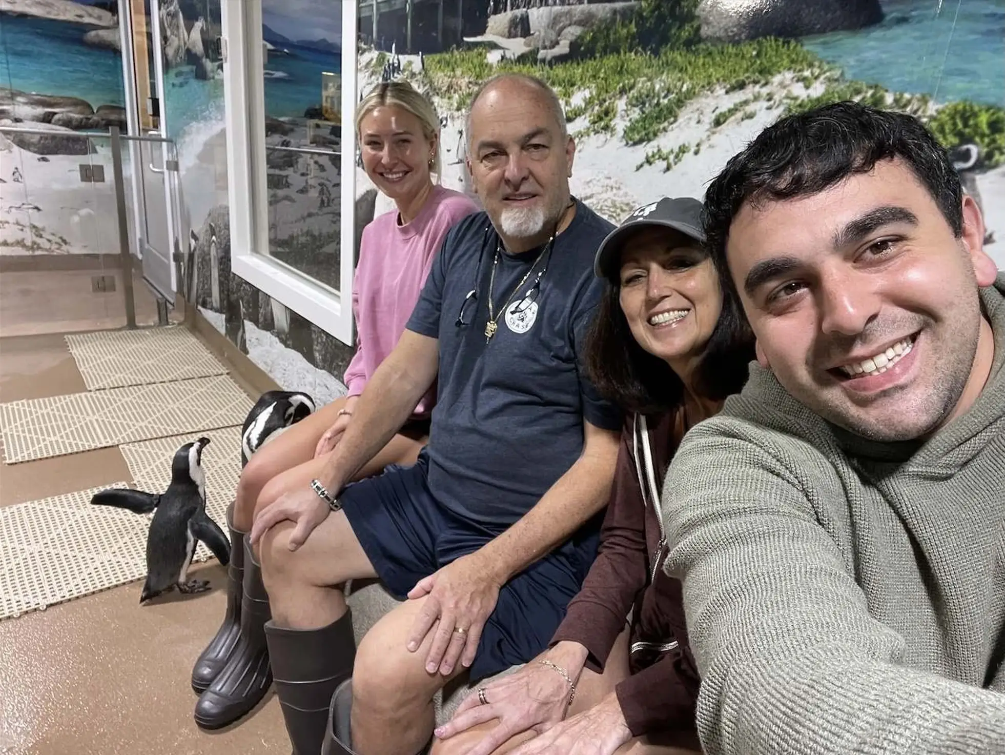 Four visitors pose for a selfie beside two penguins in an indoor coastal-themed exhibit.