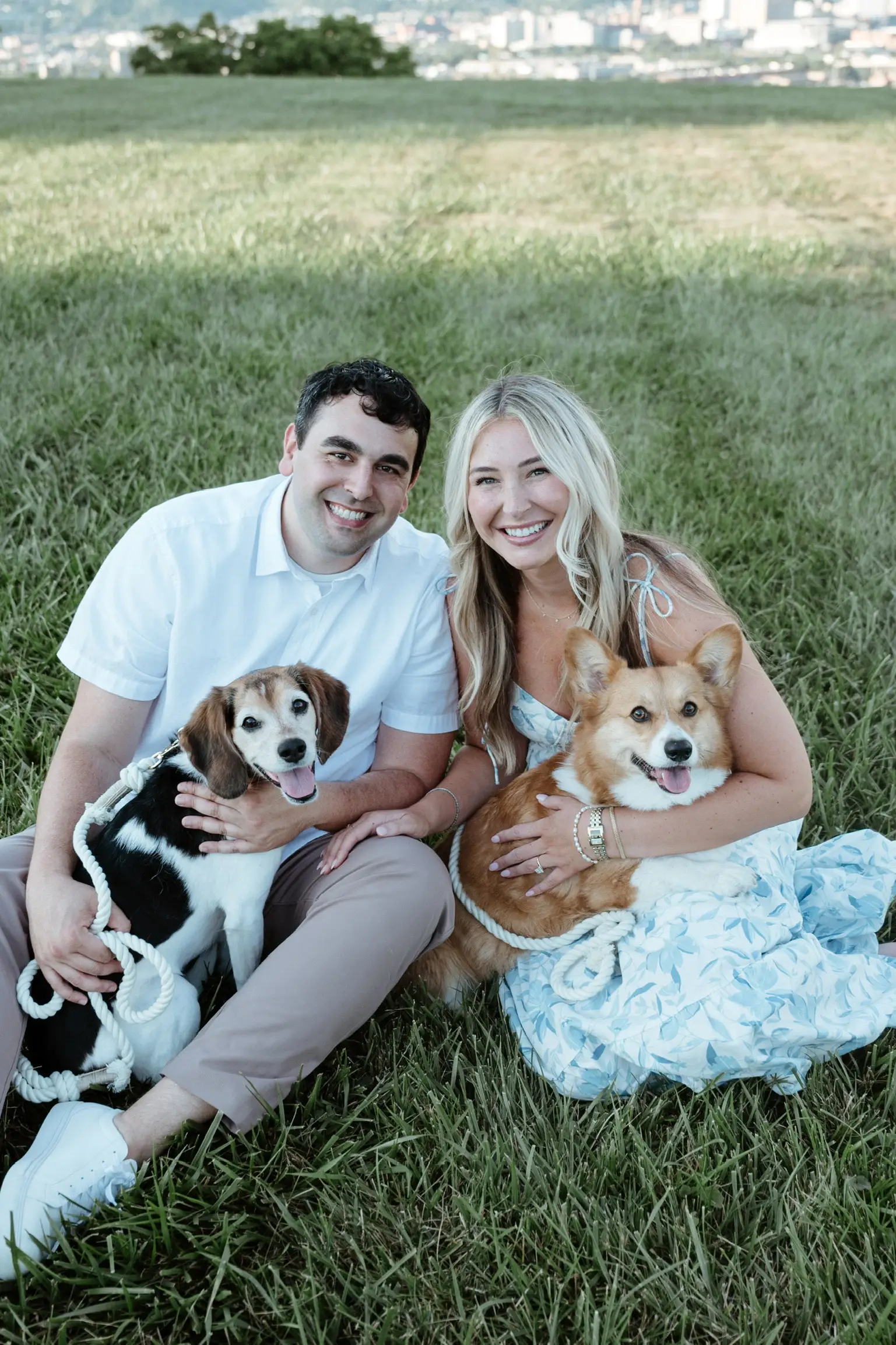 Two smiling adults sit in a grassy field holding two dogs, with a blurred city skyline in the background.