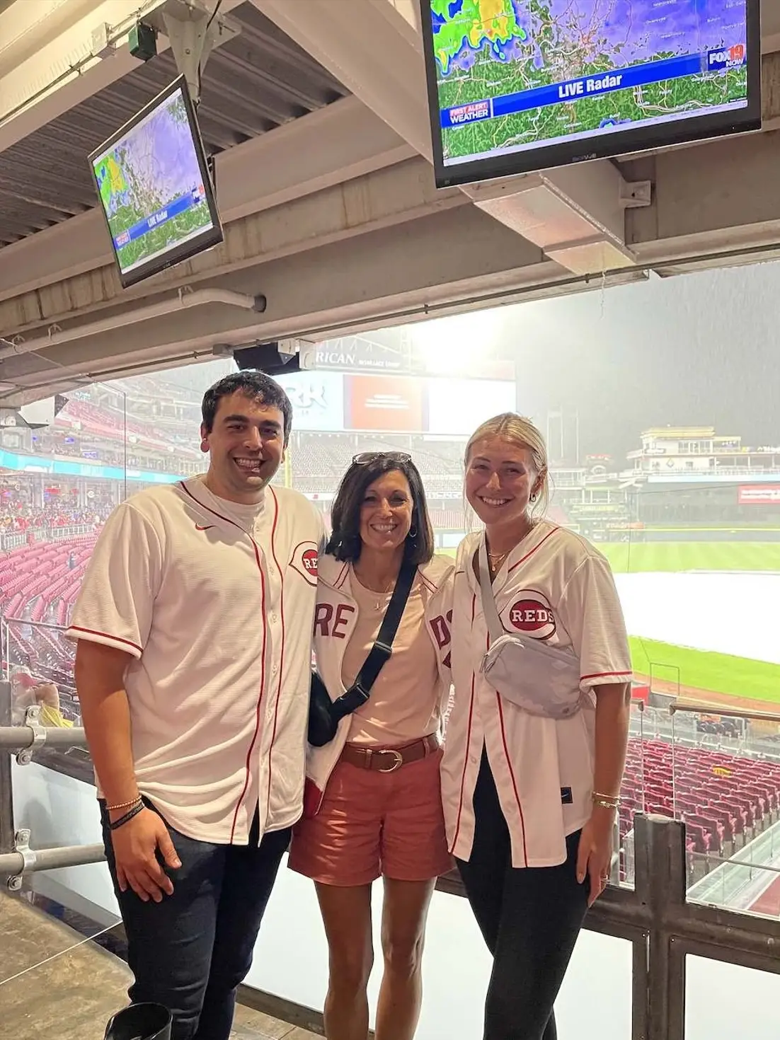 Three fans in Reds jerseys pose at a rainy baseball stadium concourse beneath live radar weather screens.