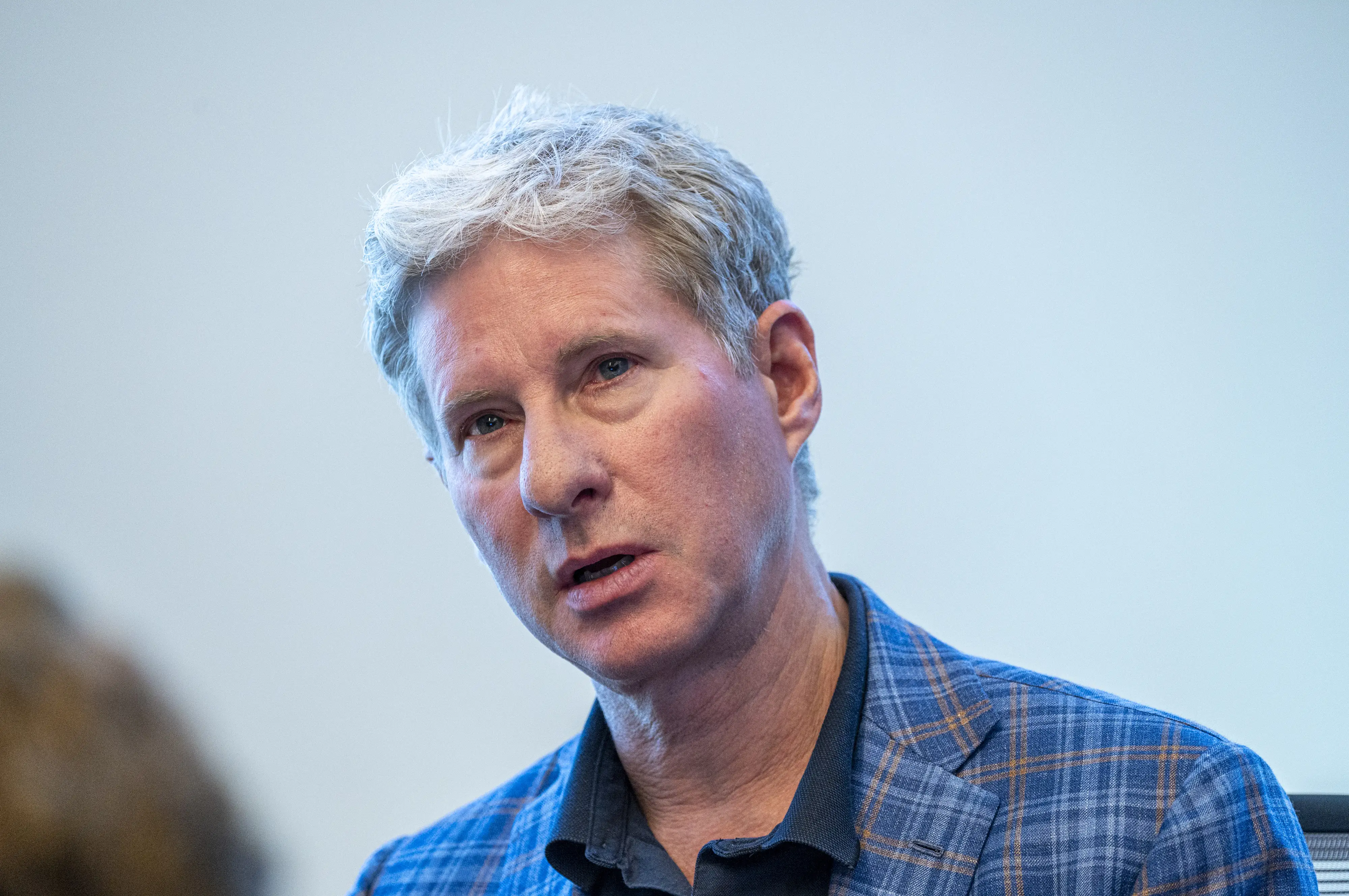 Chris Larsen in a blue plaid blazer appears in close-up against a plain light background at an indoor event.