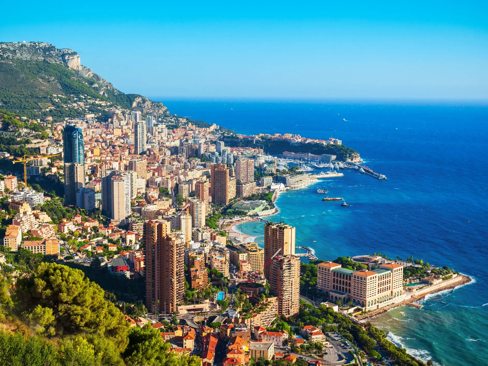 Coastal Monaco cityscape with high-rise buildings, marina, mountains, and blue Mediterranean water under a clear sky.