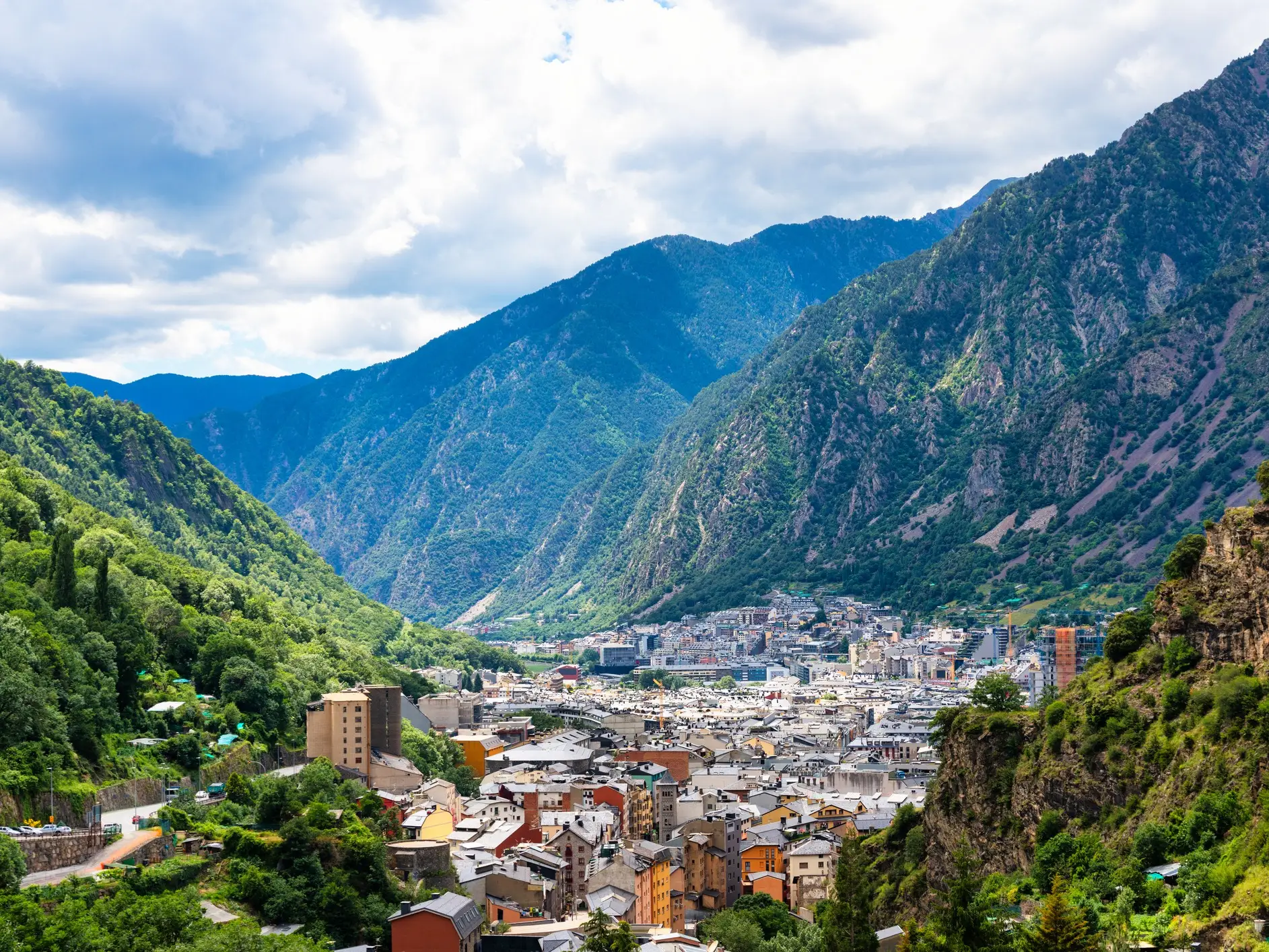 Dense town buildings fill a green mountain valley beneath steep rocky slopes and a cloudy sky.
