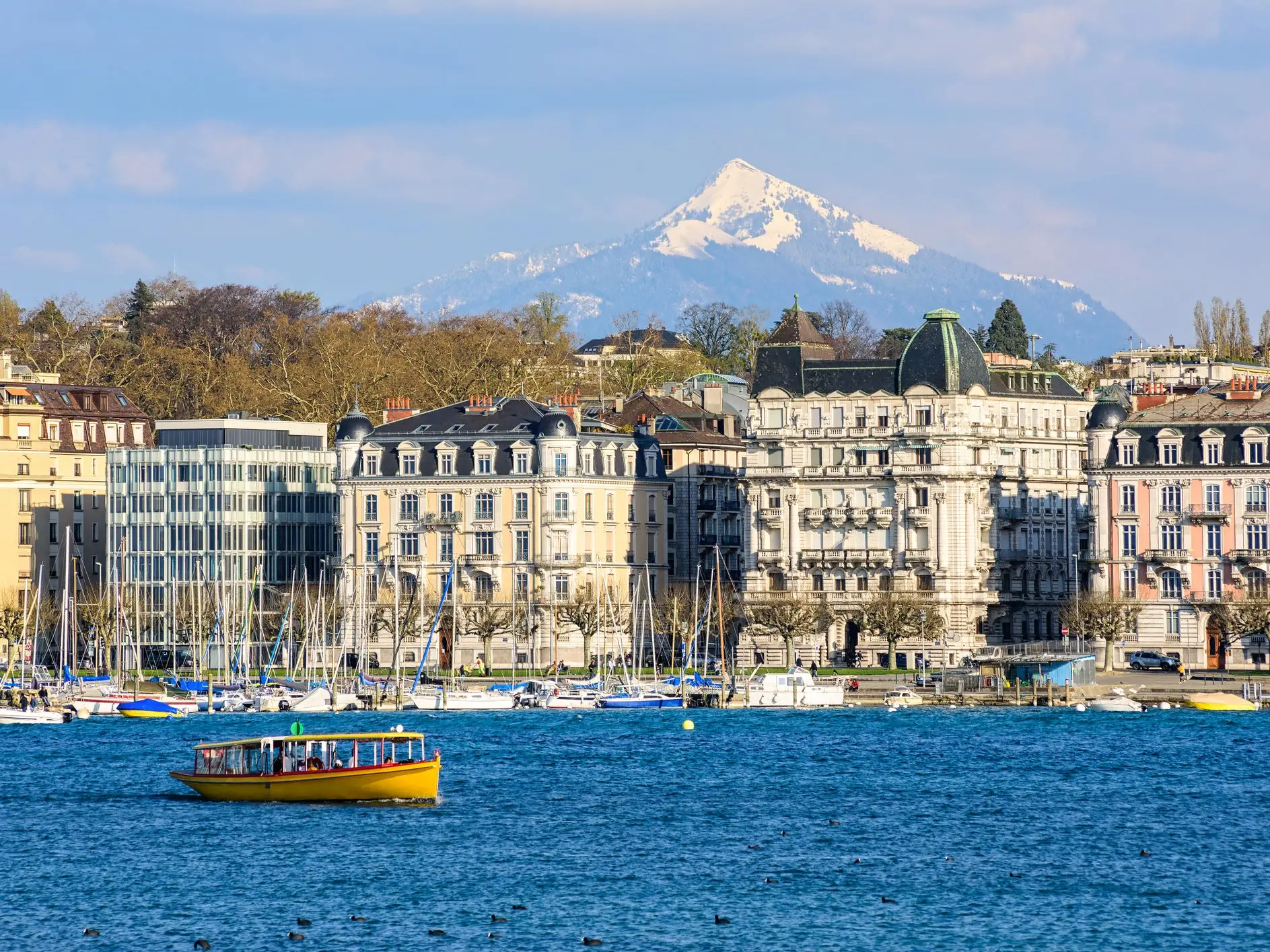 Waterfront buildings and moored sailboats line a blue lake with a yellow boat and snow-capped mountain behind.