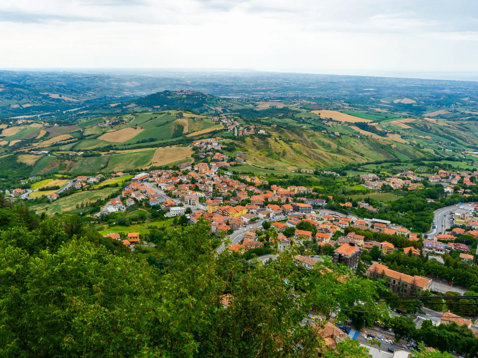 Rolling green countryside surrounds a red-roofed hillside town under a cloudy sky.