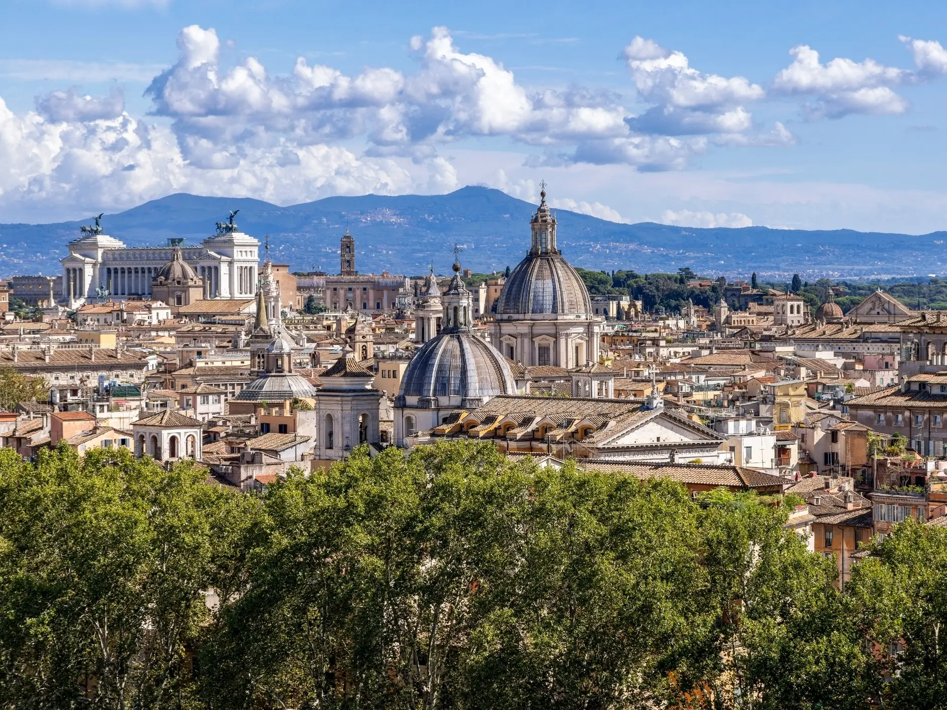 Rome skyline with historic domes, the Vittoriano monument, trees, and distant mountains under a partly cloudy sky.