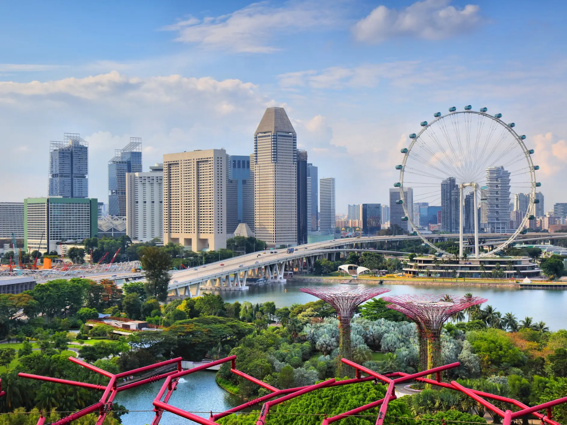 Singapore skyline with the Singapore Flyer, Marina Bay waterfront, bridge, and Gardens by the Bay greenery under a cloudy sky.
