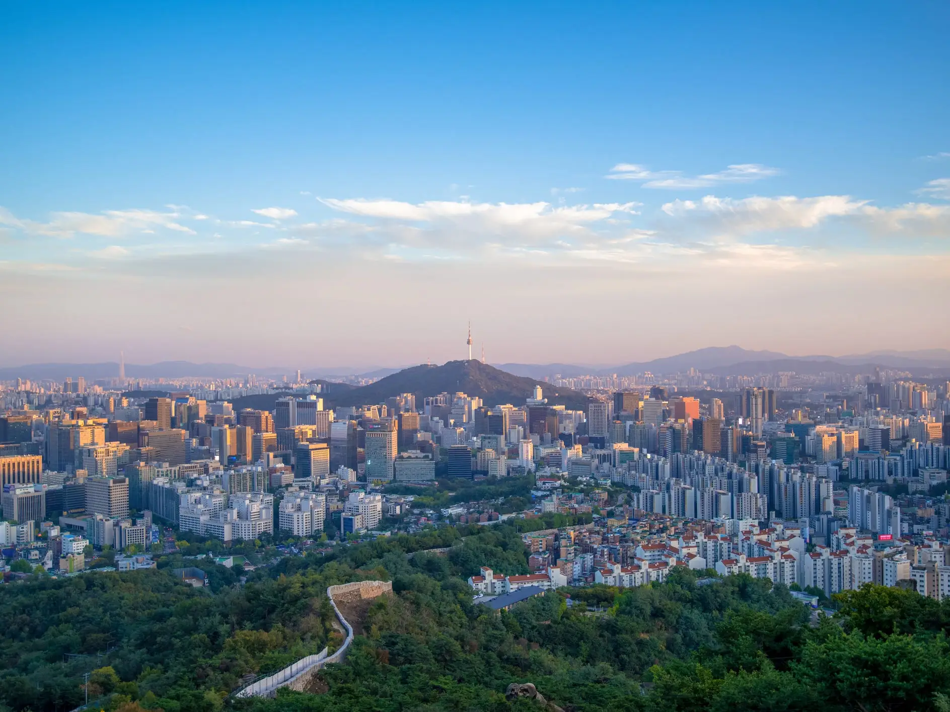 Seoul skyline at sunset with N Seoul Tower rising from a central hill above dense high-rise buildings.
