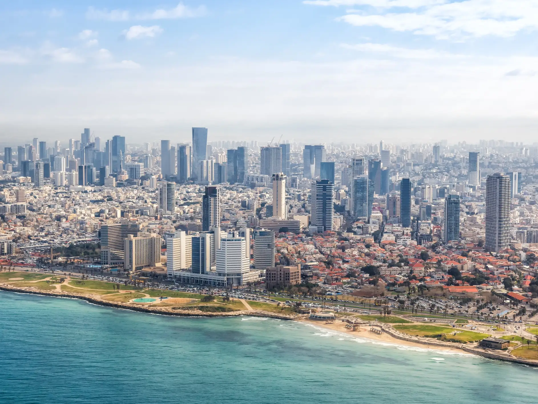 Aerial view of a coastal city skyline with high-rise buildings, beachfront parks, roads, and turquoise water.