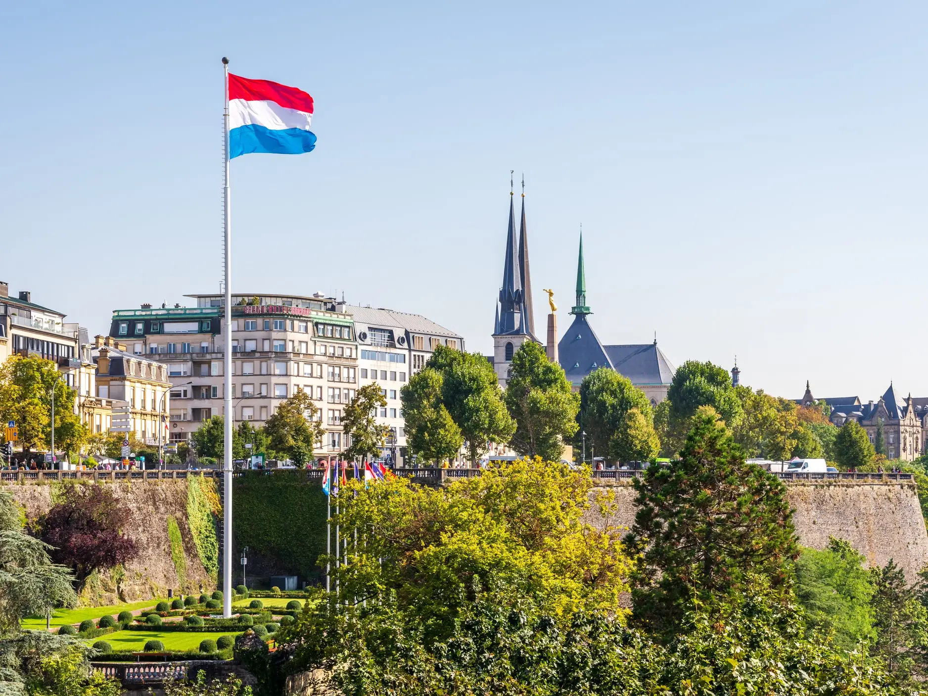 Luxembourg flag flies above a green park and historic city skyline with church spires.