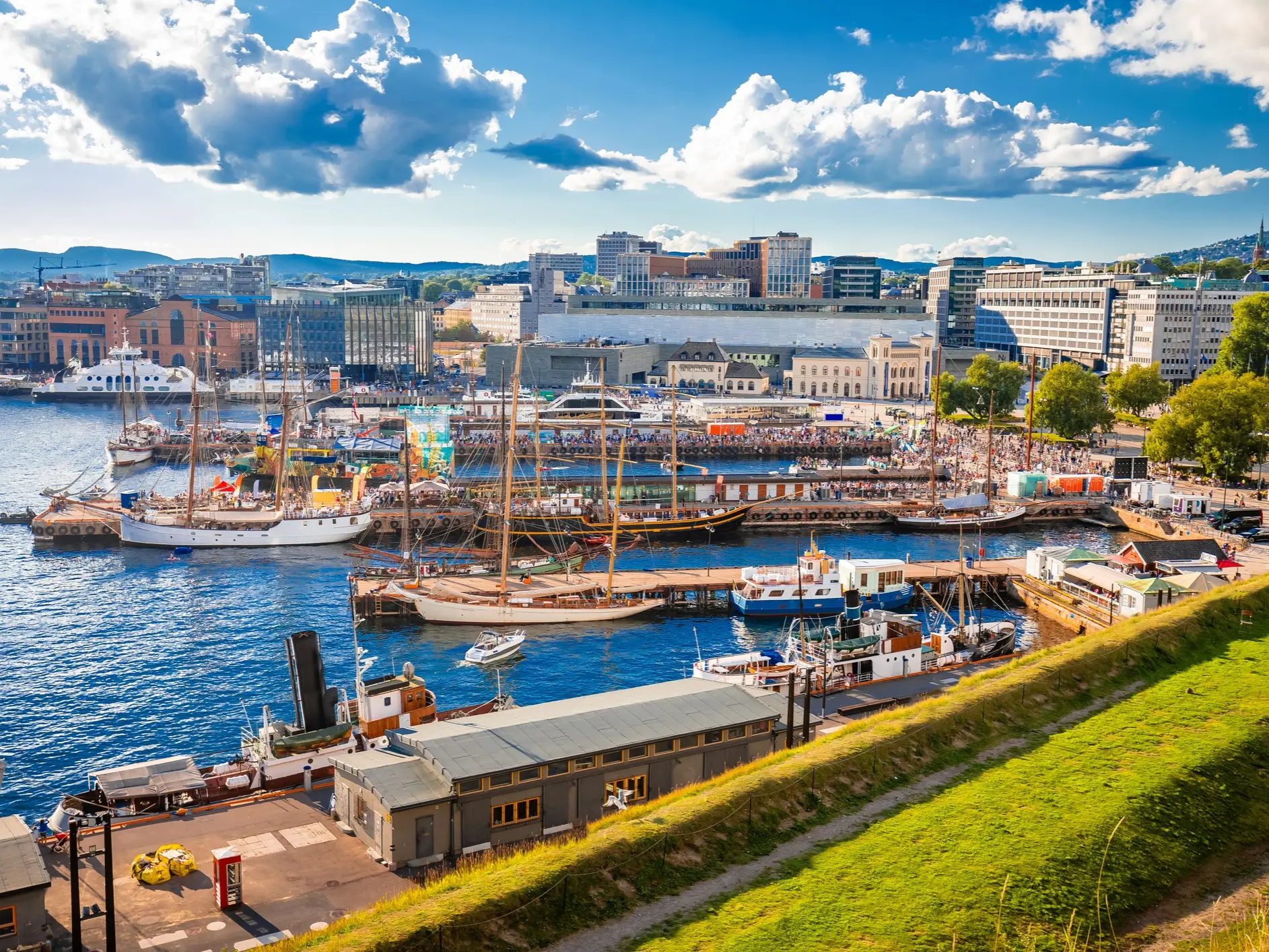 Sailboats and crowds fill Oslo's sunny waterfront harbor beneath a blue sky and city skyline.