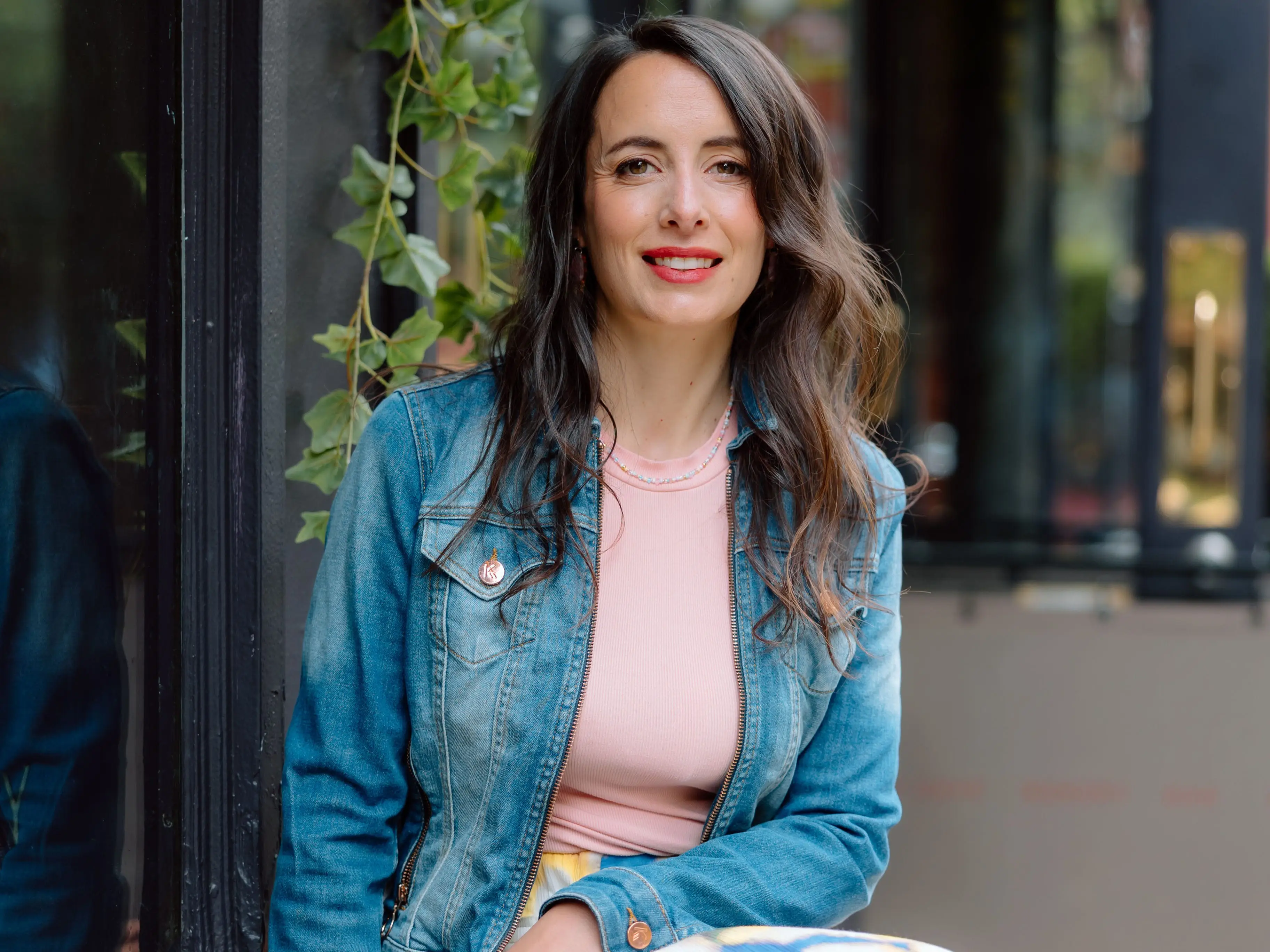 Julie Chapon in a denim jacket sits on a wooden bench beside a window with hanging greenery.