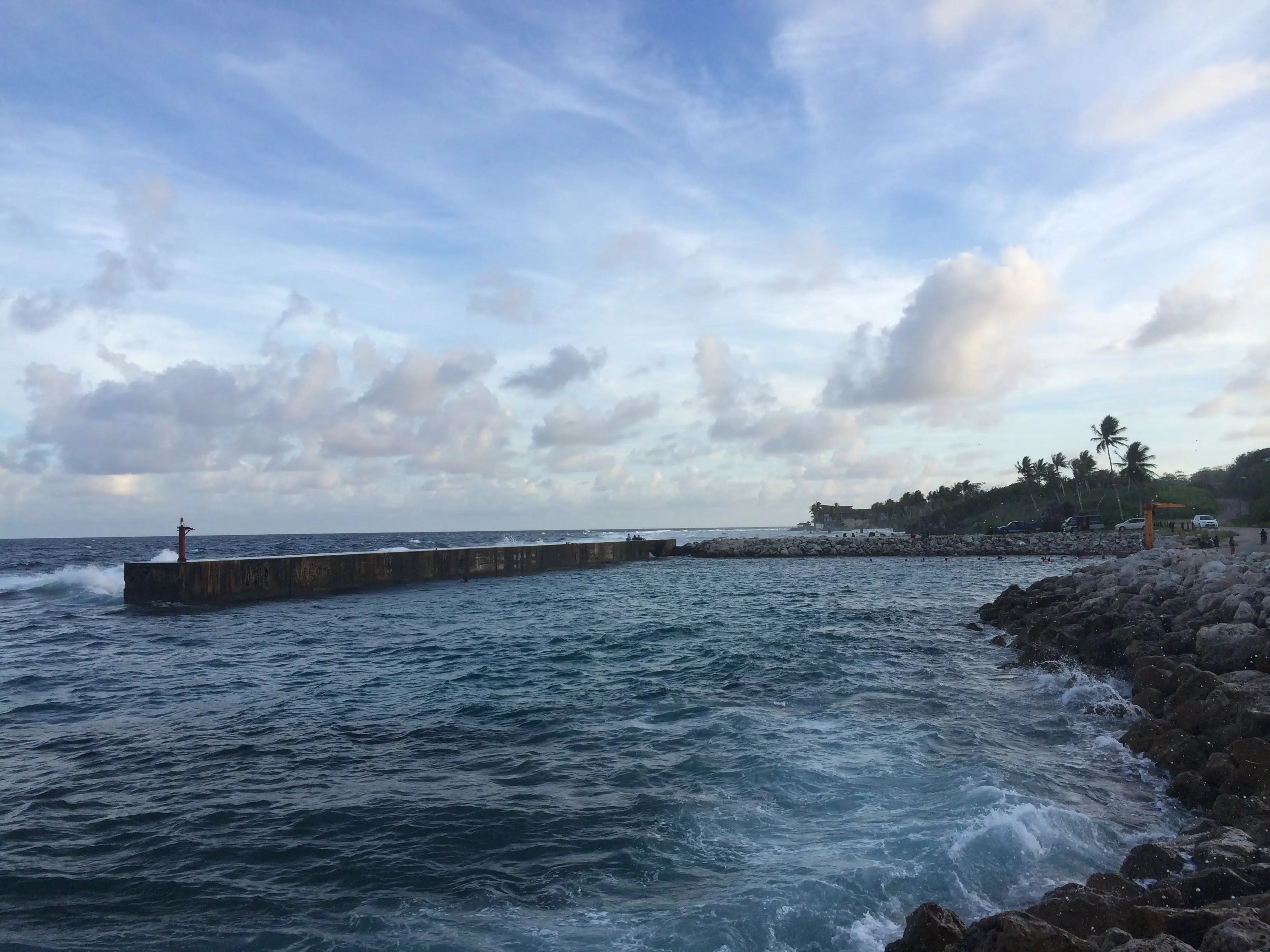 Ocean waves break against a harbor breakwater and rocky shoreline beneath a cloudy blue sky.