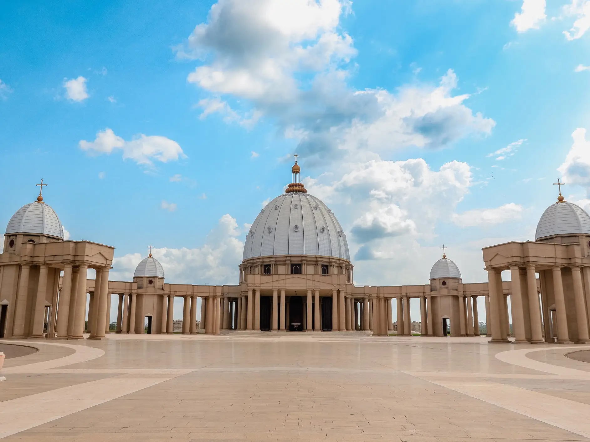 Domed basilica with colonnades stands across a wide paved plaza beneath a partly cloudy blue sky.
