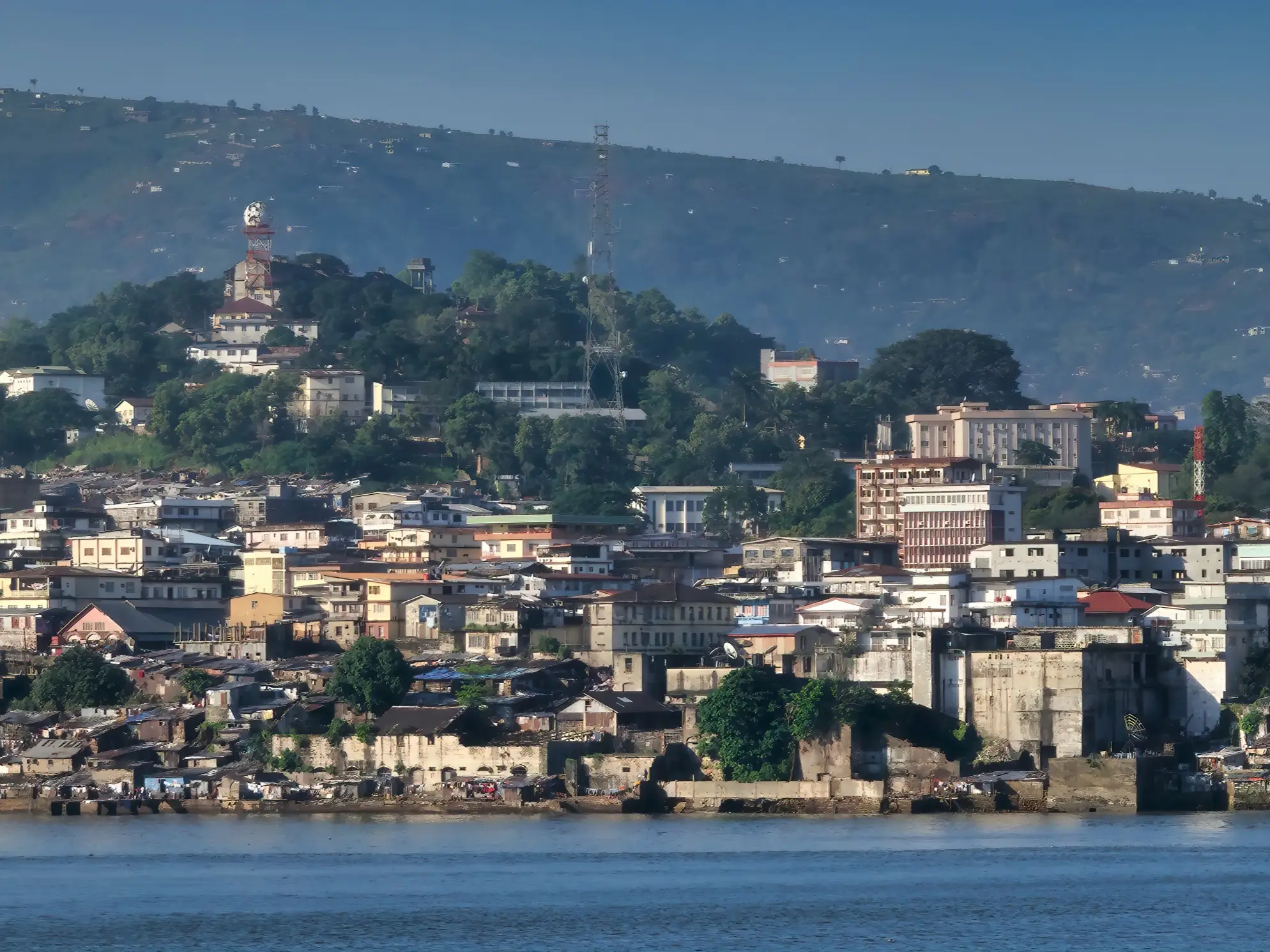 Dense waterfront city buildings sit below green hills with communication towers under a clear blue sky.