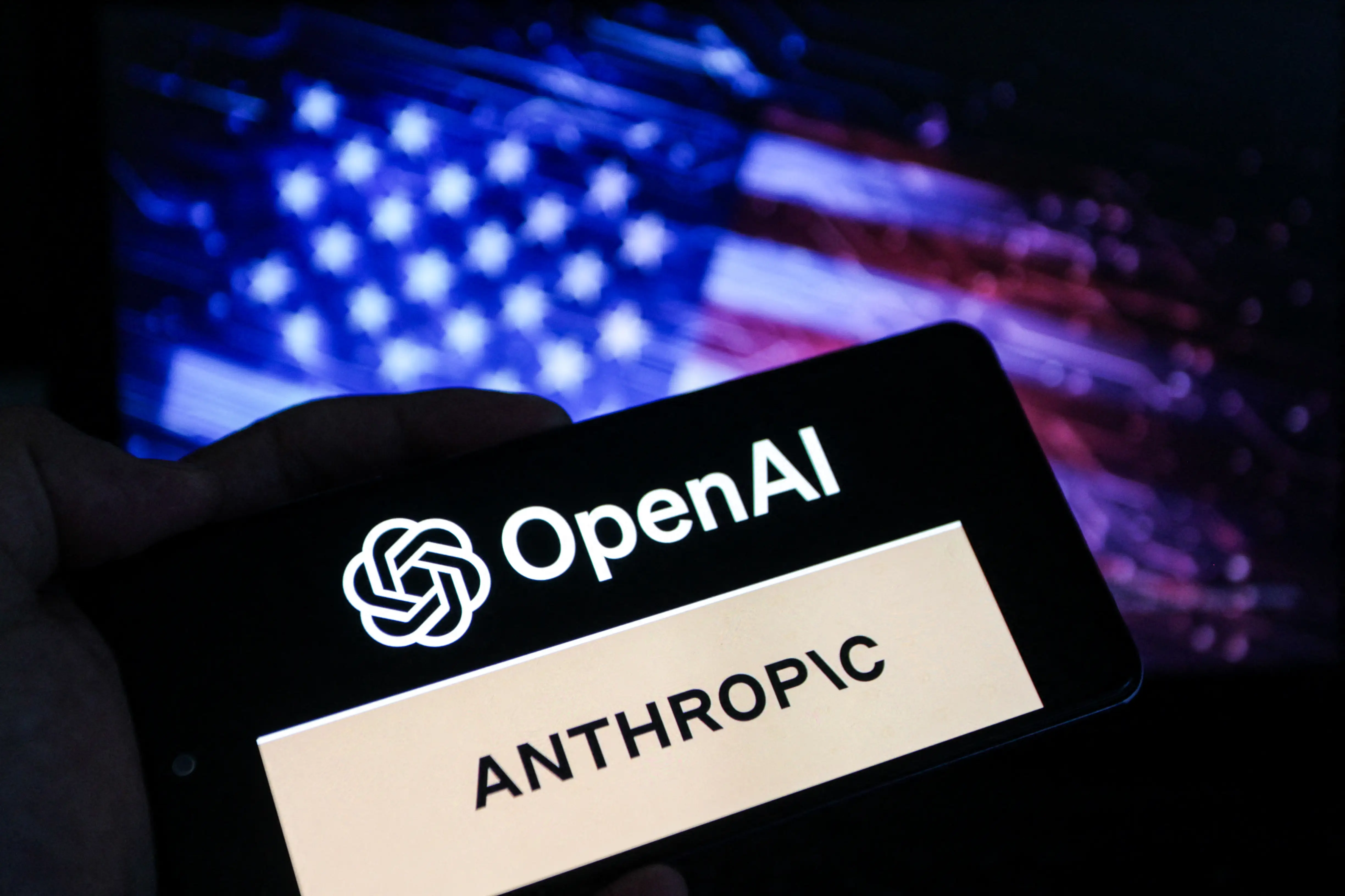 A smartphone screen displays the logos of OpenAI and Anthropic against a blurred backdrop of the American flag