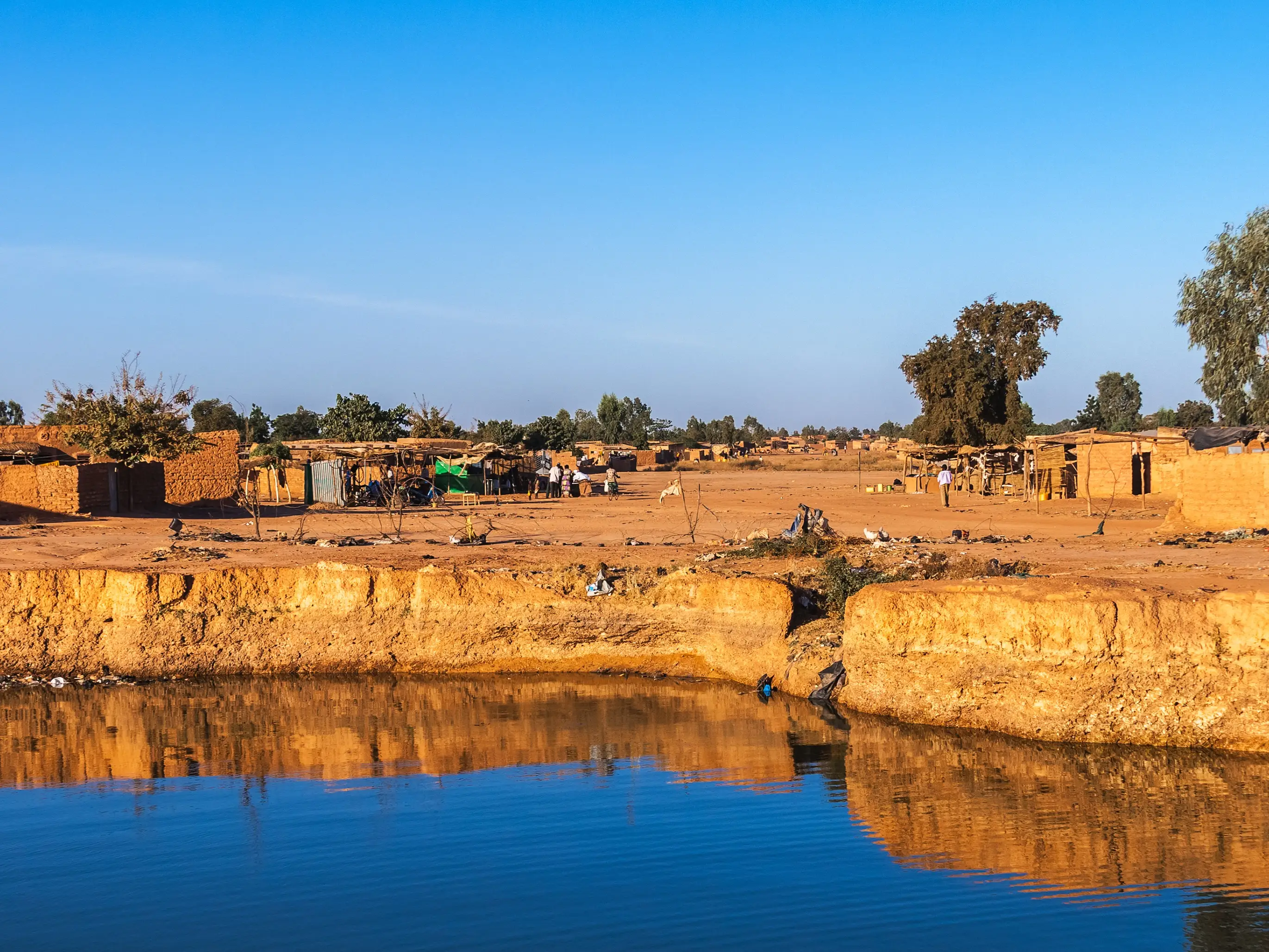 Mud-brick buildings and small shelters line a calm waterway under a clear blue sky.