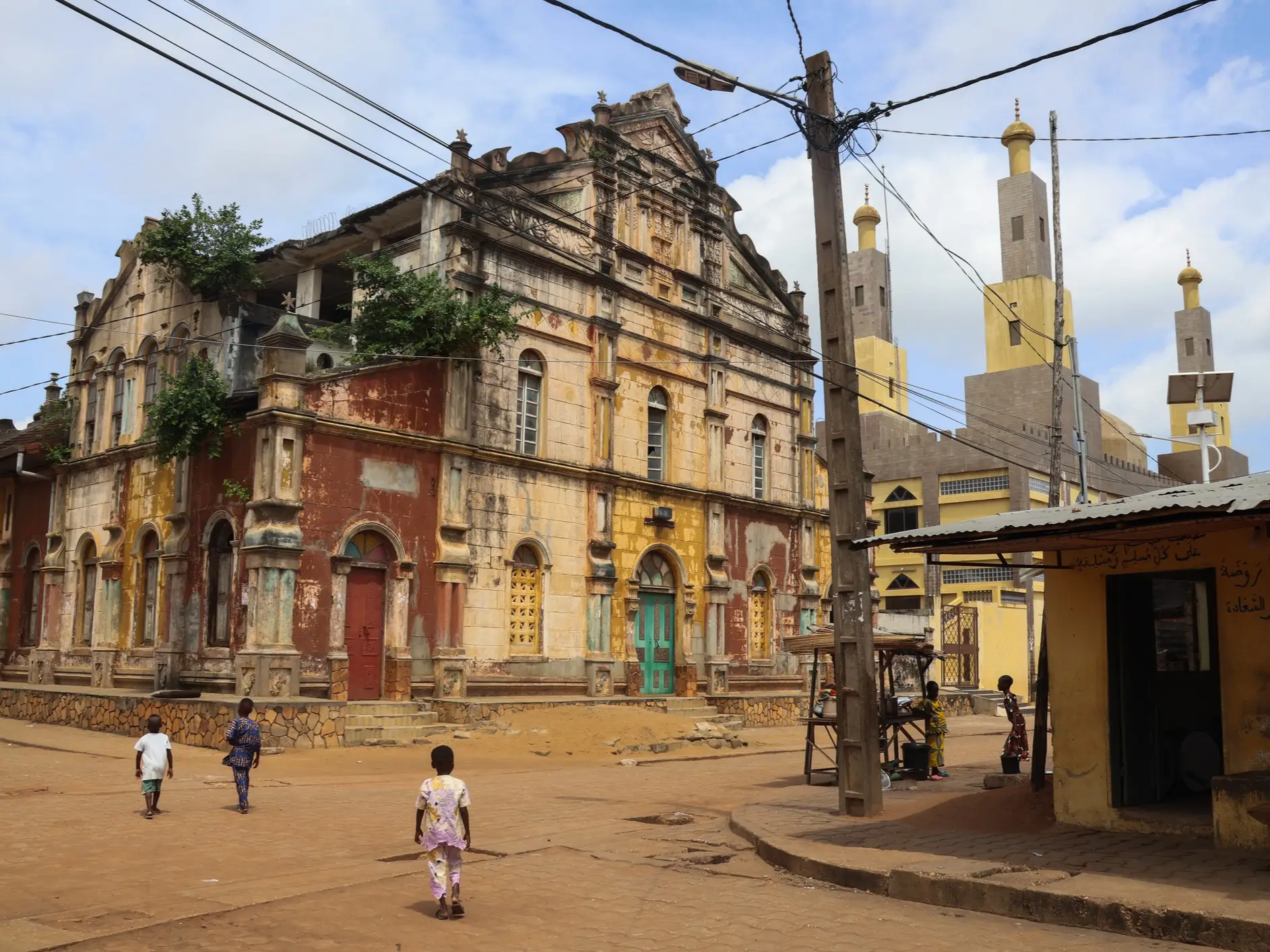 Weathered historic building stands beside a mosque with minarets on a quiet street crossed by overhead wires.