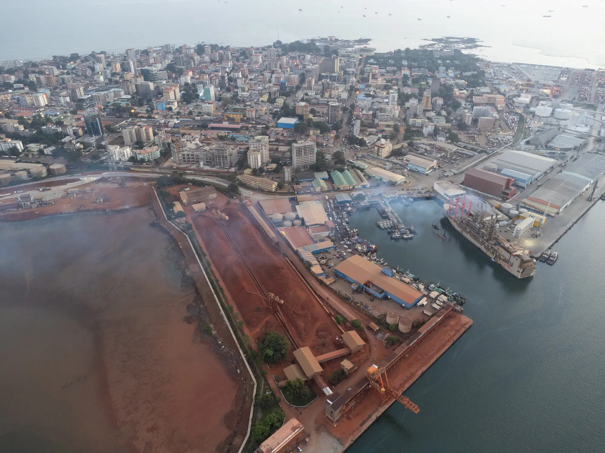 Aerial view of a coastal city with port facilities, docks, warehouses, cranes, and vessels along the waterfront.