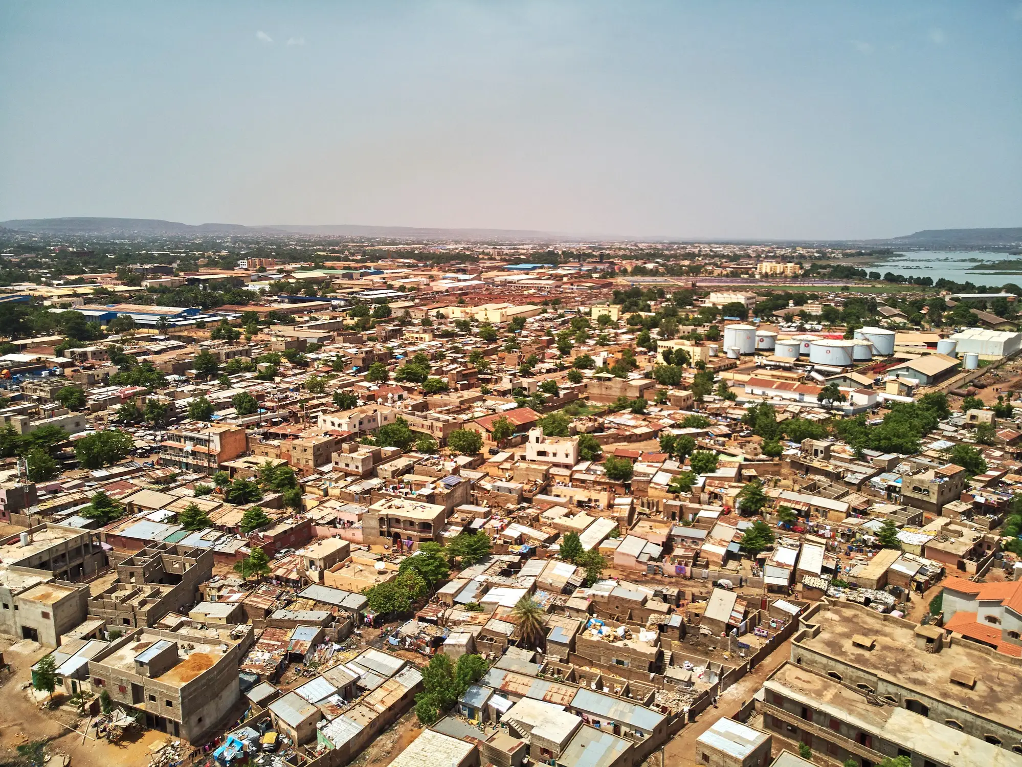 Aerial view of a dense low-rise city district with industrial storage tanks and water visible in the distance.