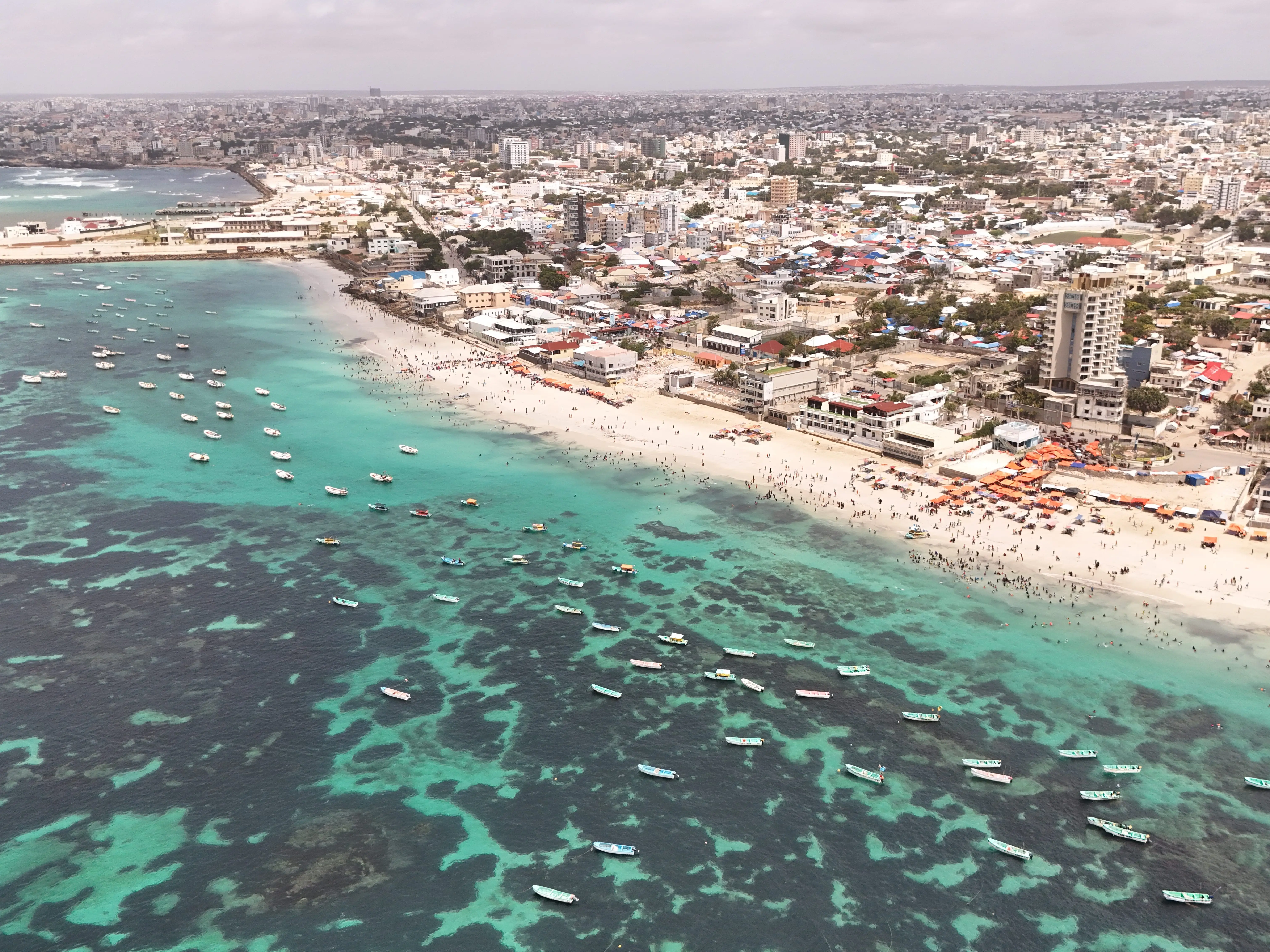 Aerial view of a crowded urban beach with turquoise coastal water, anchored boats, and dense city buildings.