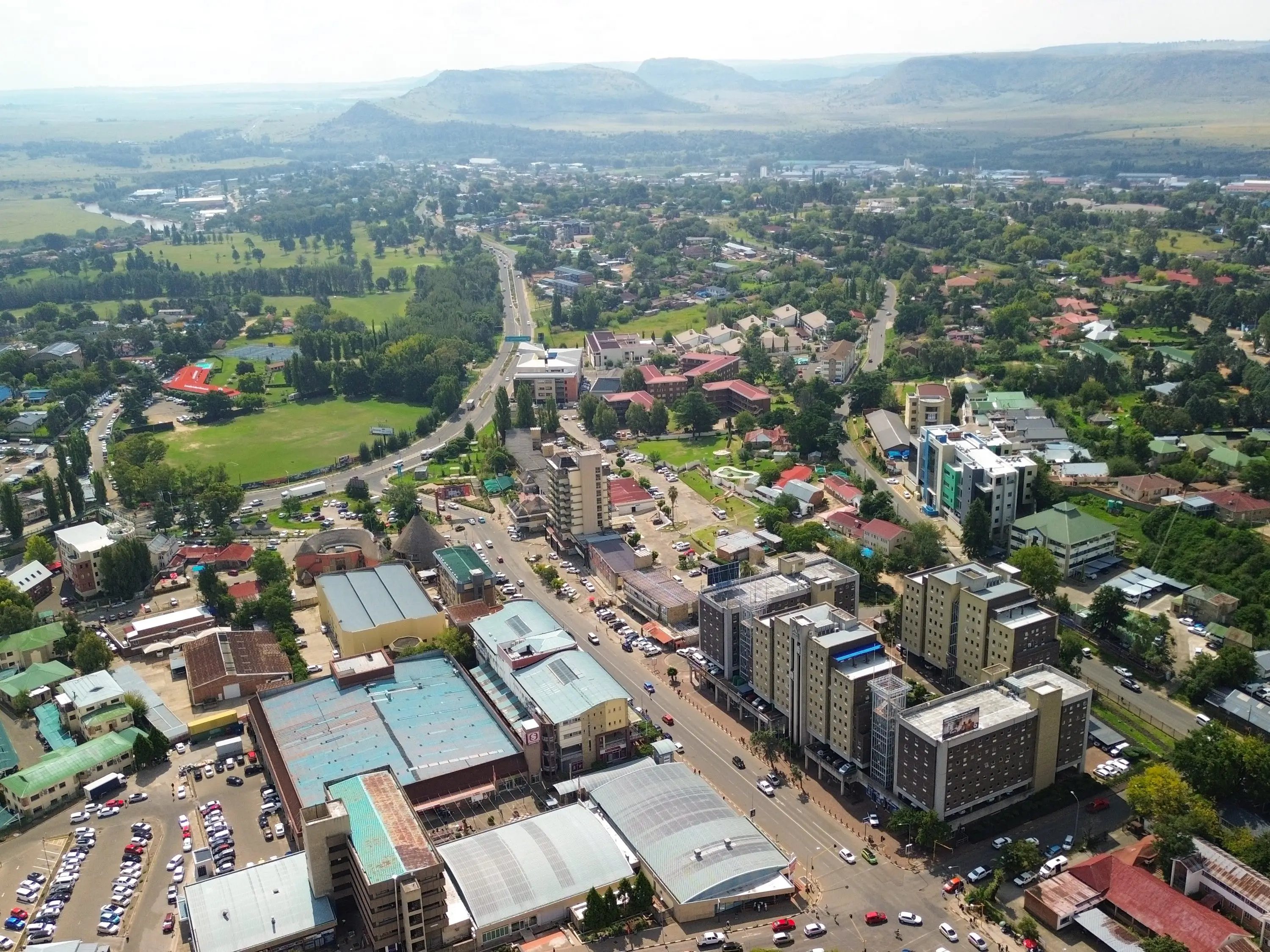 Aerial view of a town with roads, buildings, green fields, and distant hills under a hazy sky.