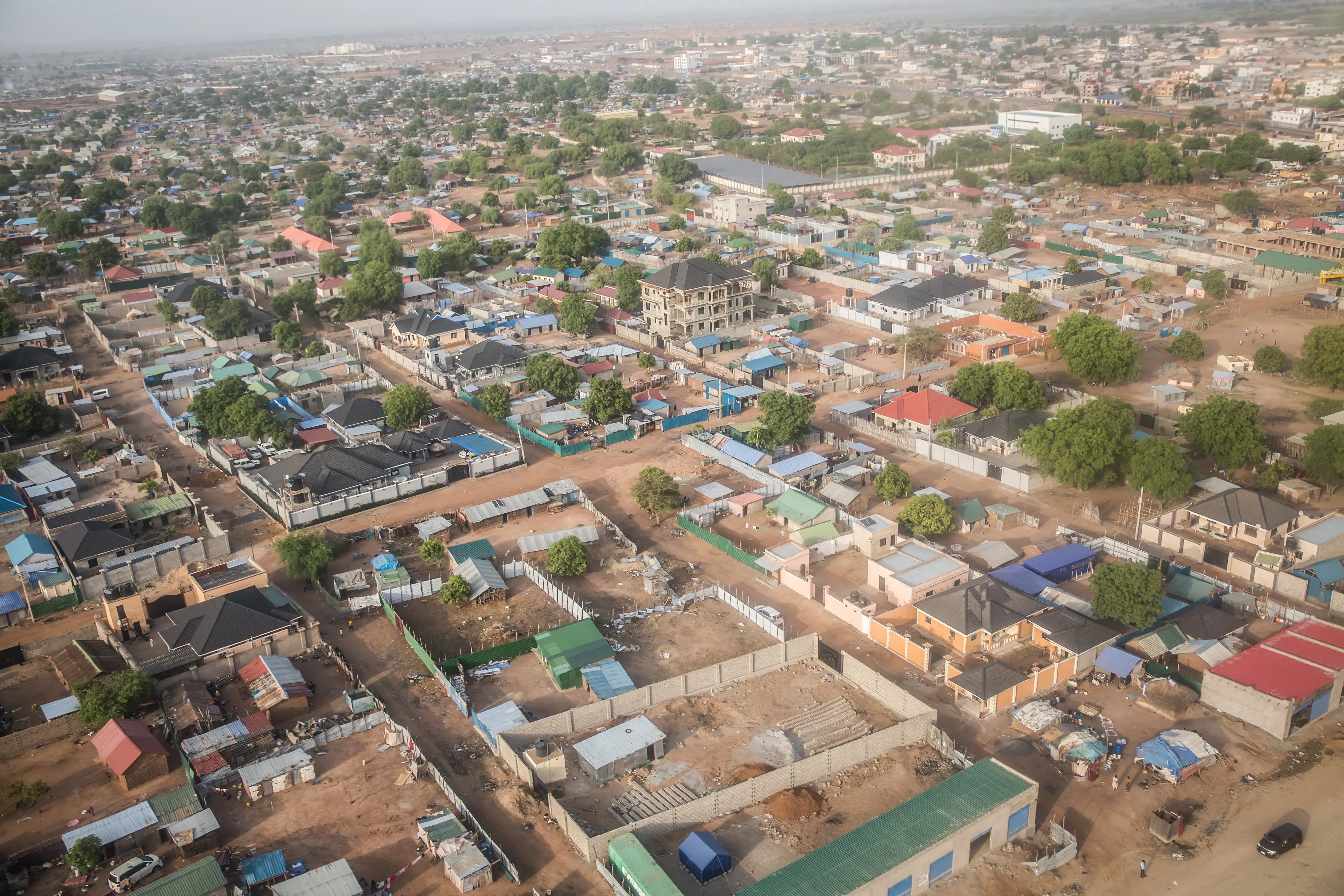 Aerial view of a sprawling low-rise urban neighborhood with dusty roads, walled compounds, trees, and scattered larger buildings.