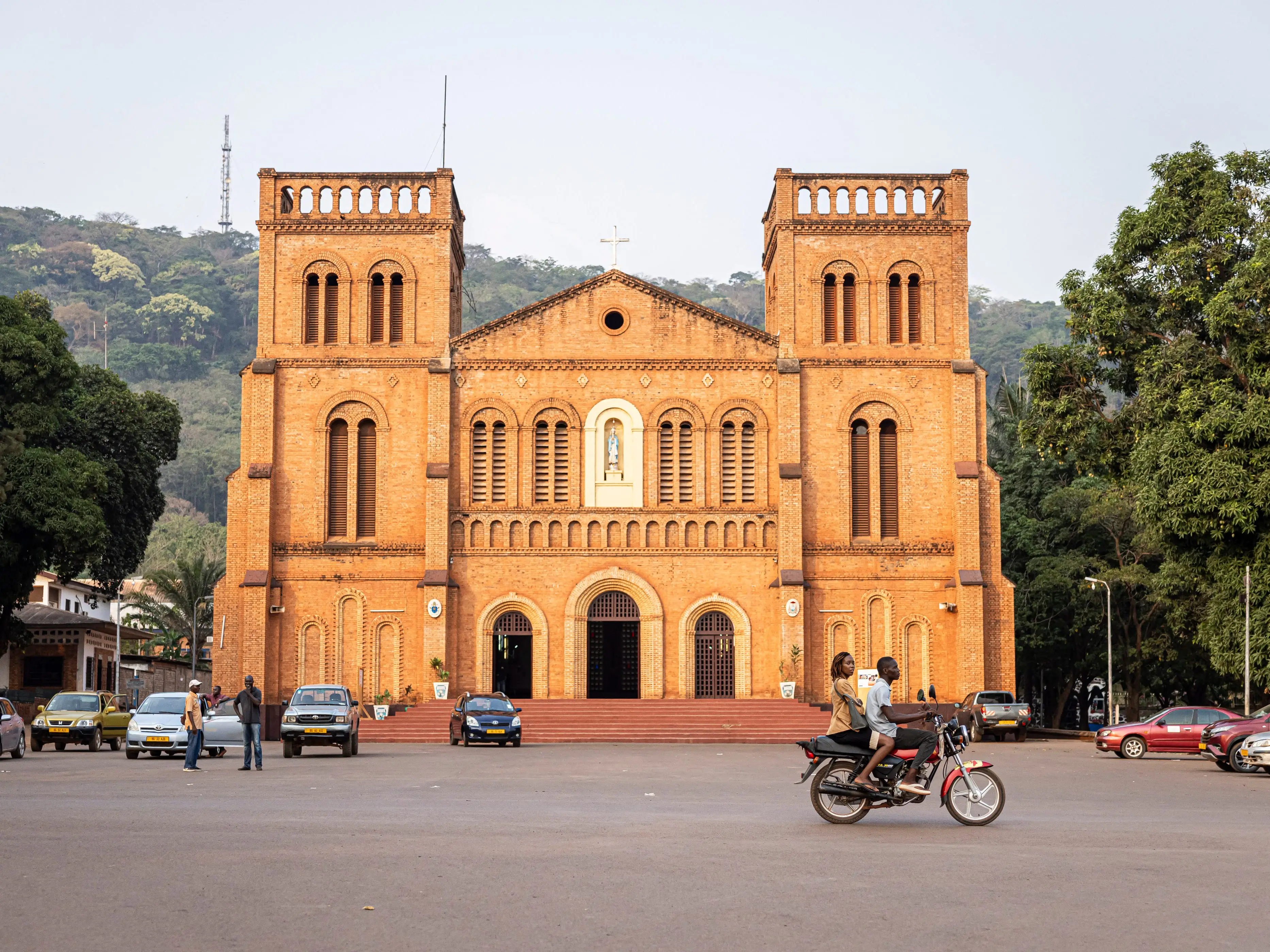 Brick church with twin towers faces a plaza with cars, pedestrians, and a motorcycle in front.