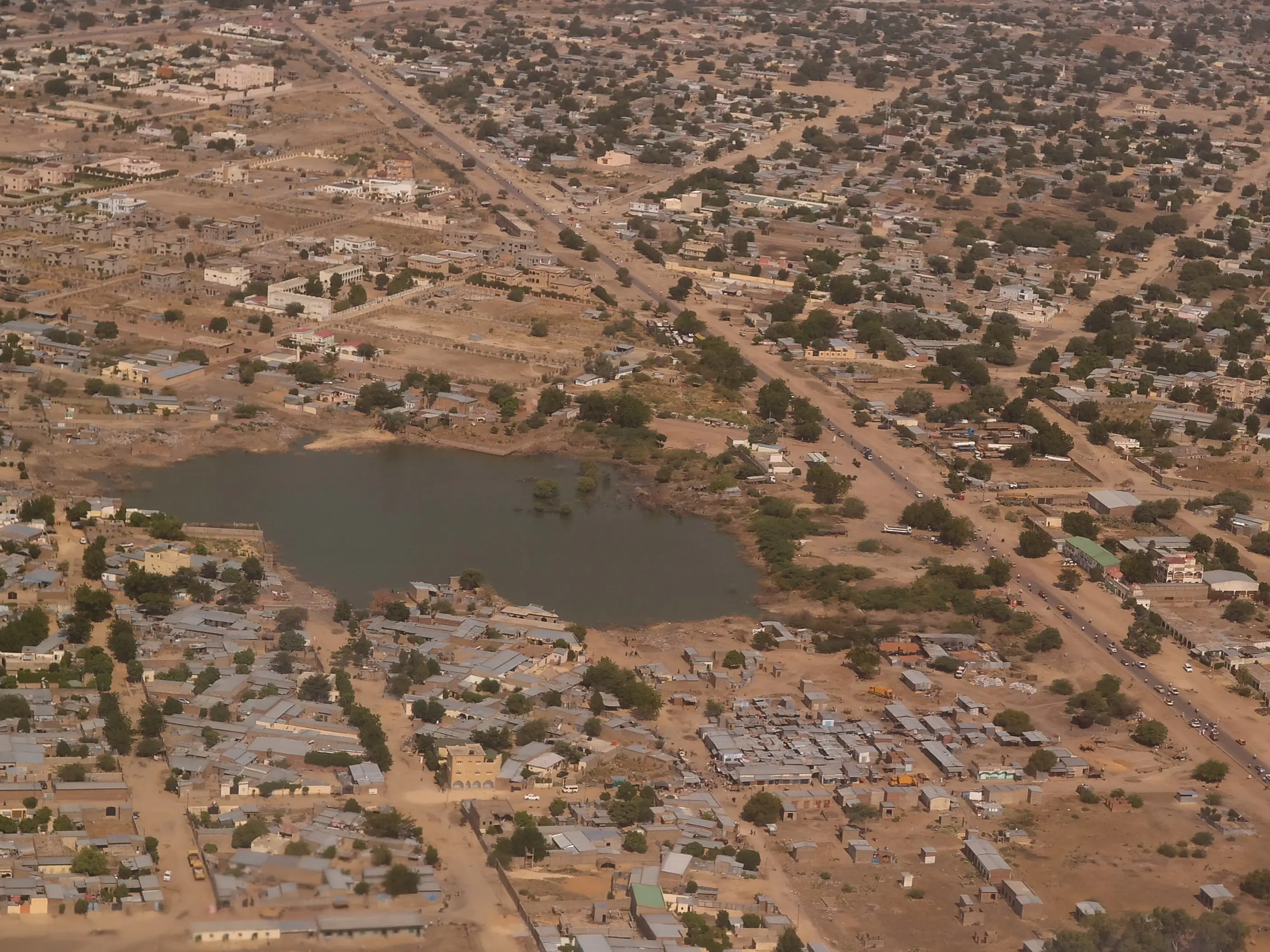 Aerial view of an arid town with dense buildings, roads, scattered trees, and a large dark pond.