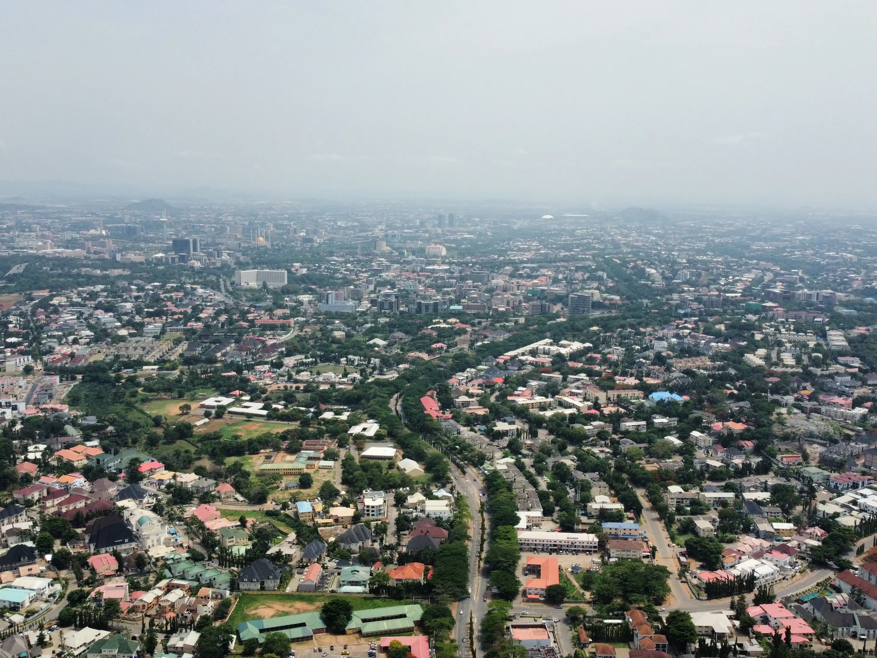 Aerial view of a sprawling city with residential neighborhoods, roads, greenery, and hazy skyline in the distance.