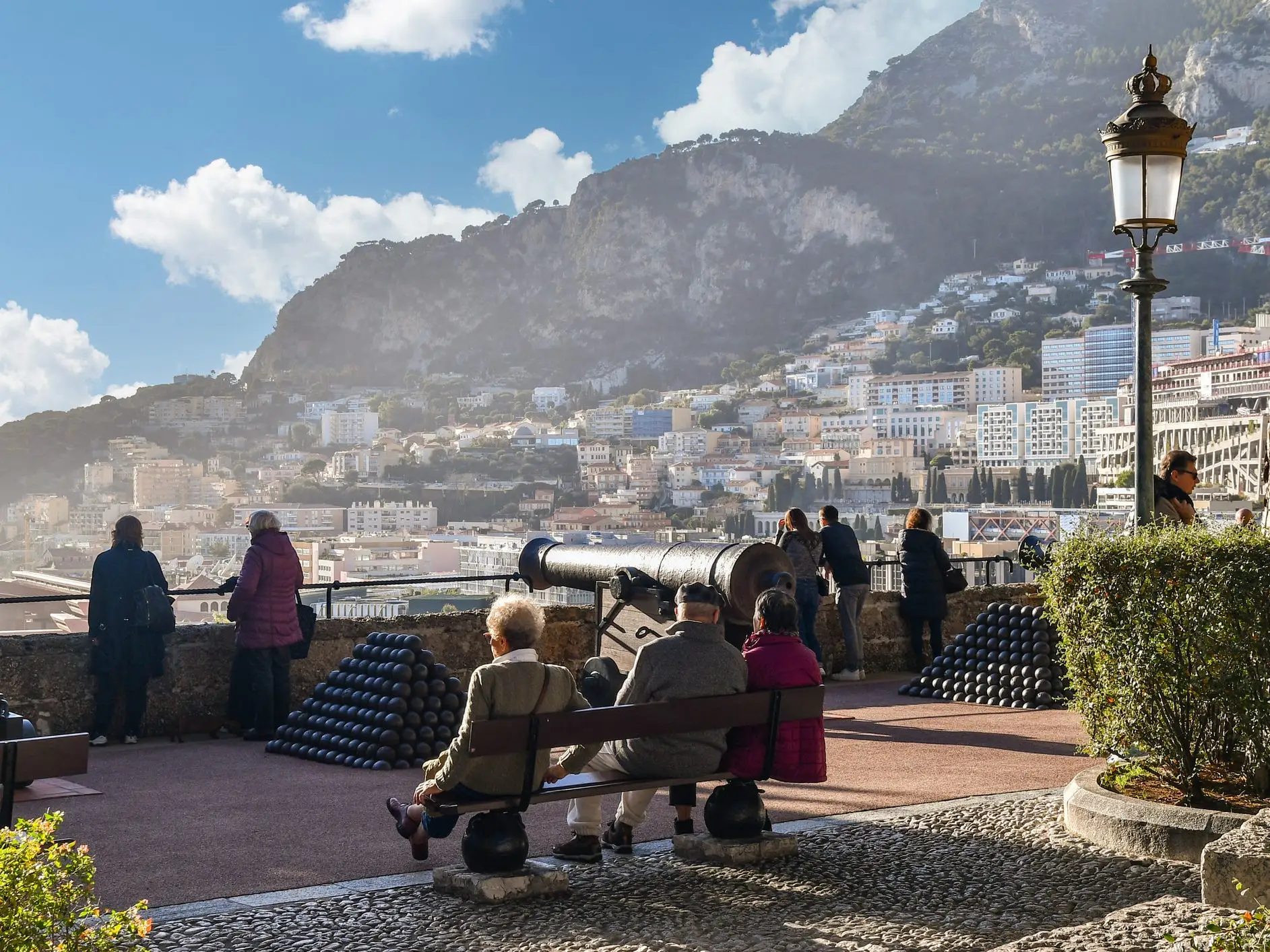 Visitors sit and stand among historic cannons overlooking a sunlit hillside city and harbor.
