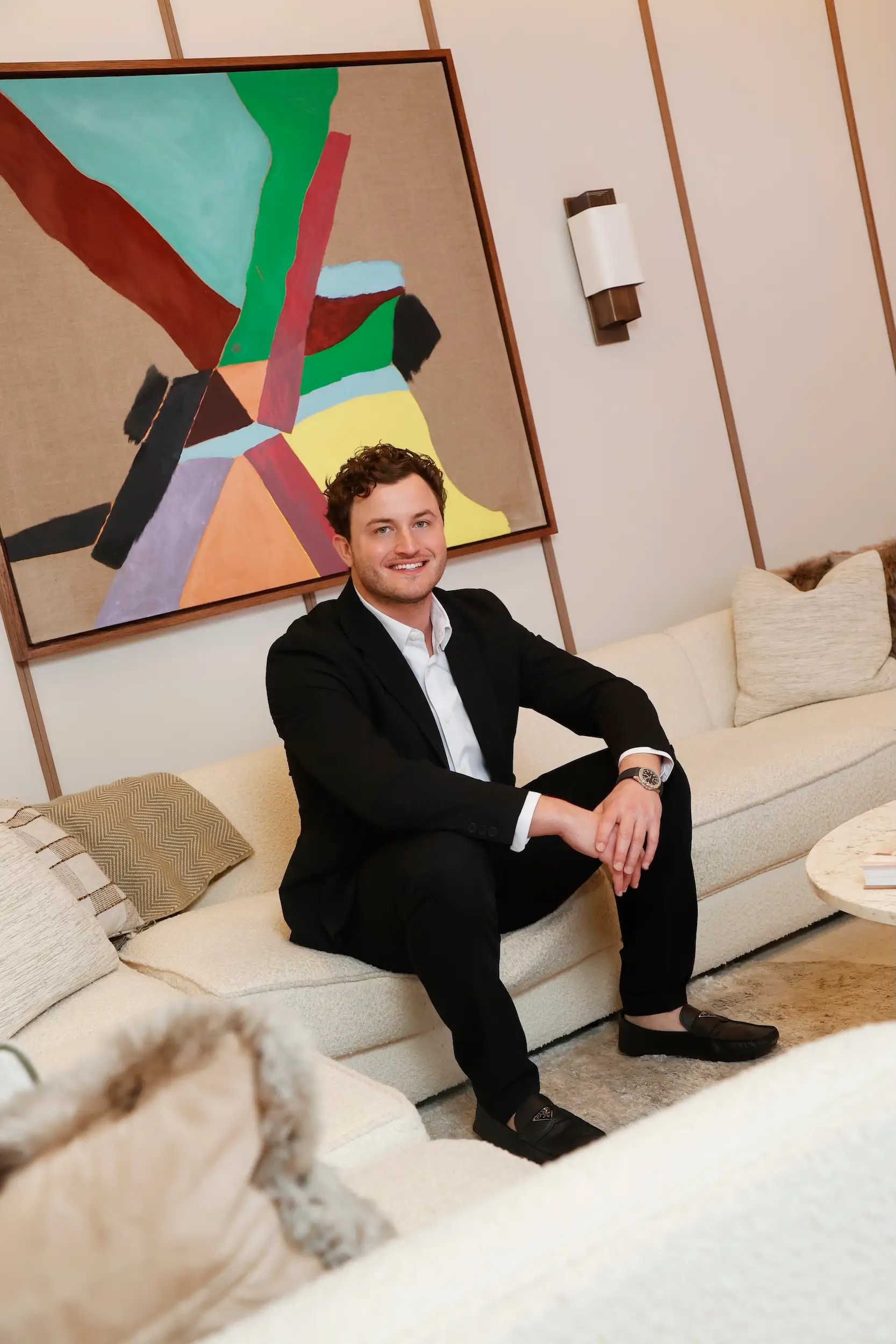 A suited person sits on a cream sofa beneath a large colorful abstract painting in a modern interior.
