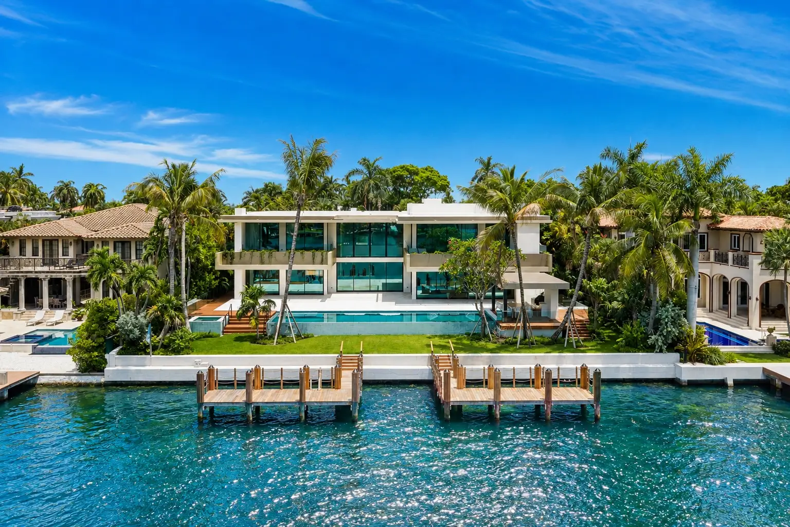 Modern waterfront mansion with palm trees, swimming pool, lawn, and private docks along bright blue water.