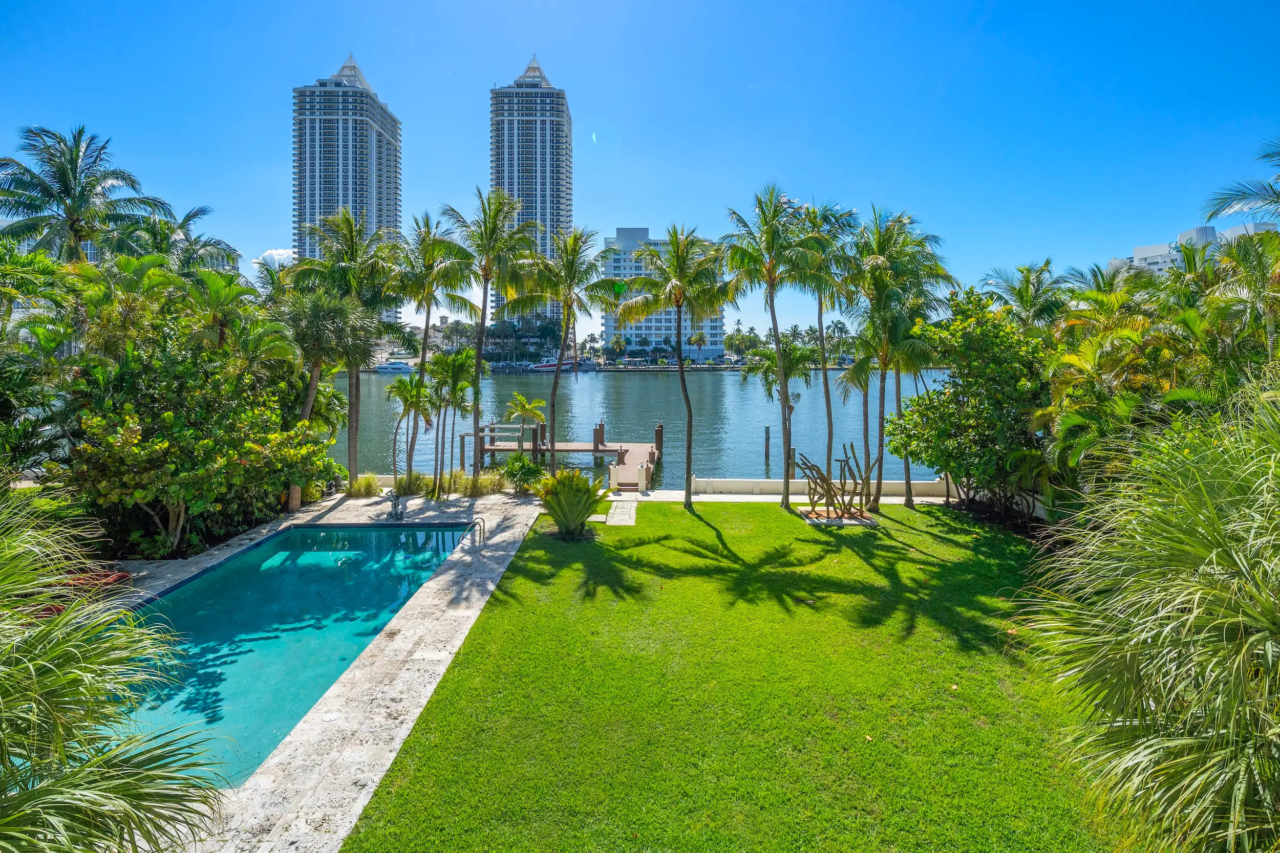 Waterfront backyard with a swimming pool, palm trees, lawn, dock, and high-rise buildings across the water.