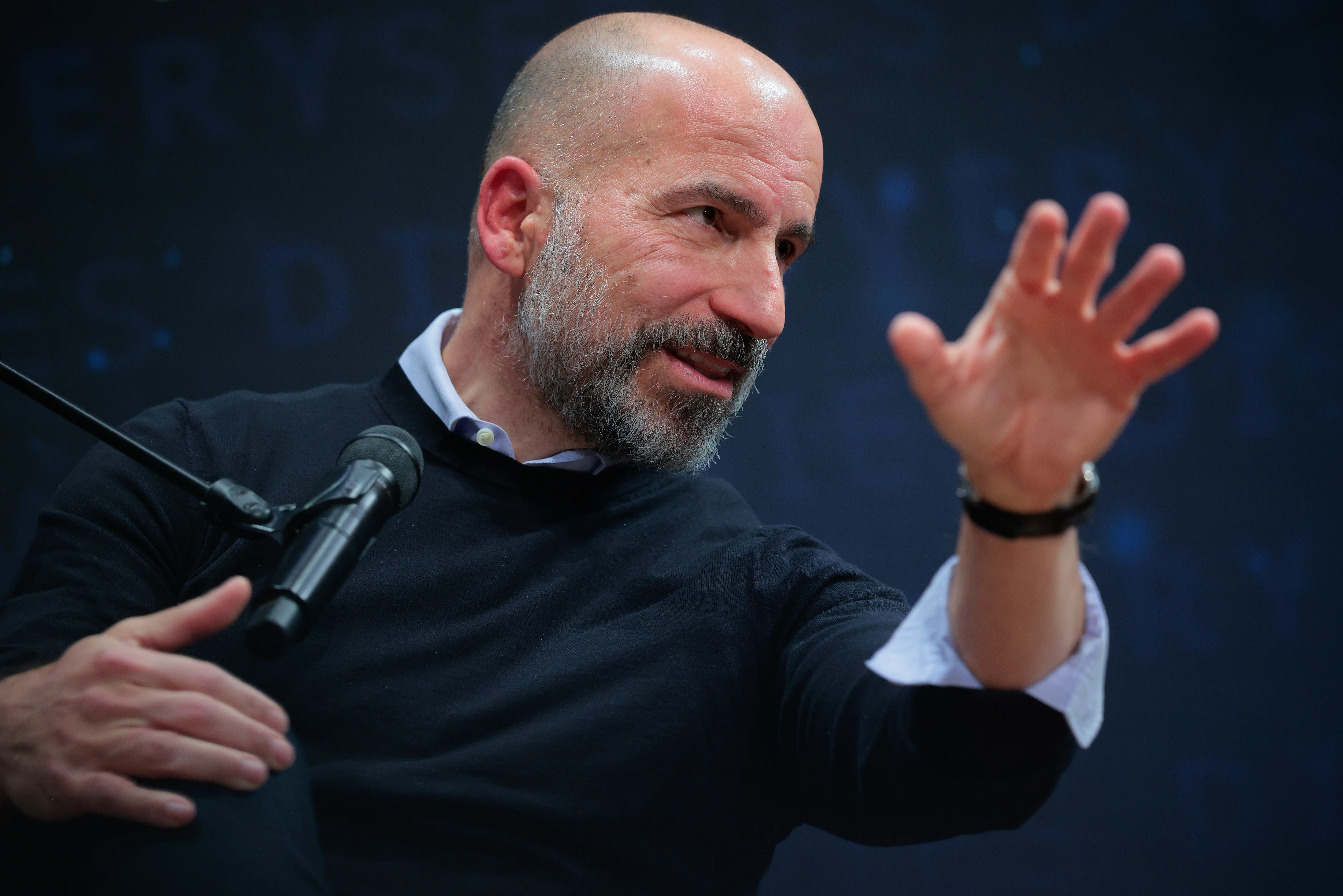 Dara Khosrowshahi gestures while speaking into a microphone on a dark conference stage.