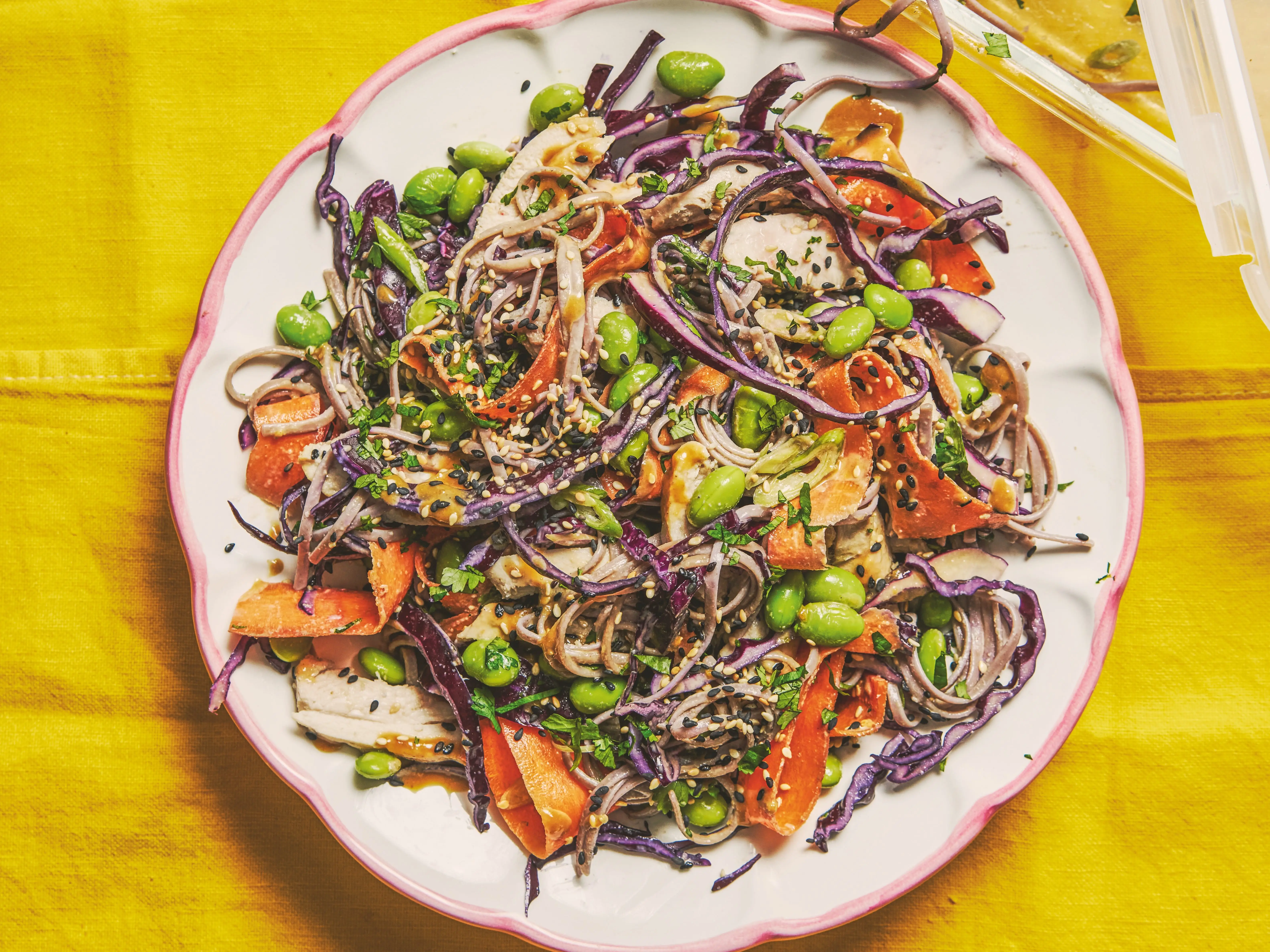 Colorful noodle salad with edamame, shredded vegetables, sesame seeds, and dressing on a yellow tablecloth.