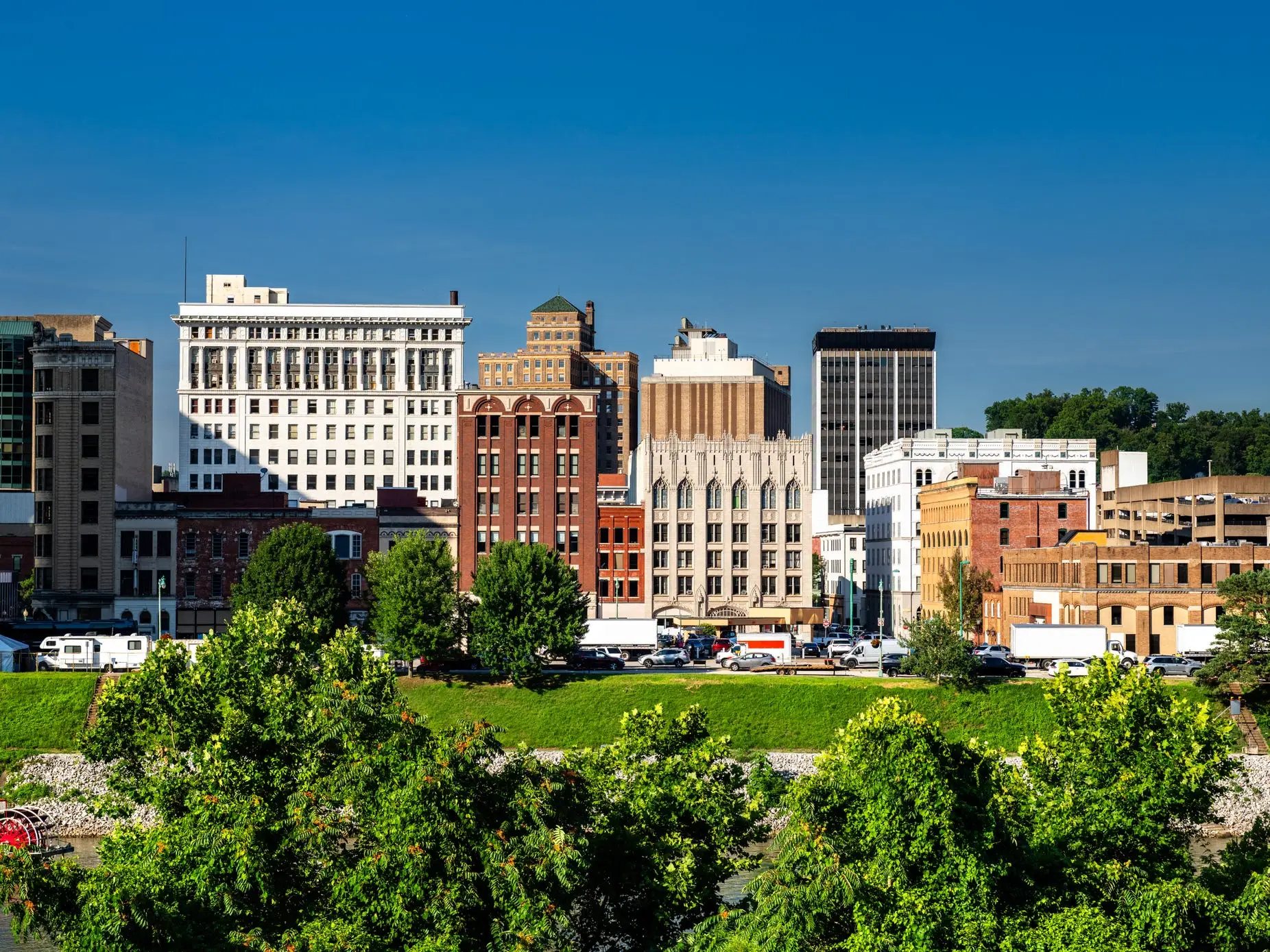 Downtown Charleston skyline rises behind trees and a riverfront on a clear sunny day.