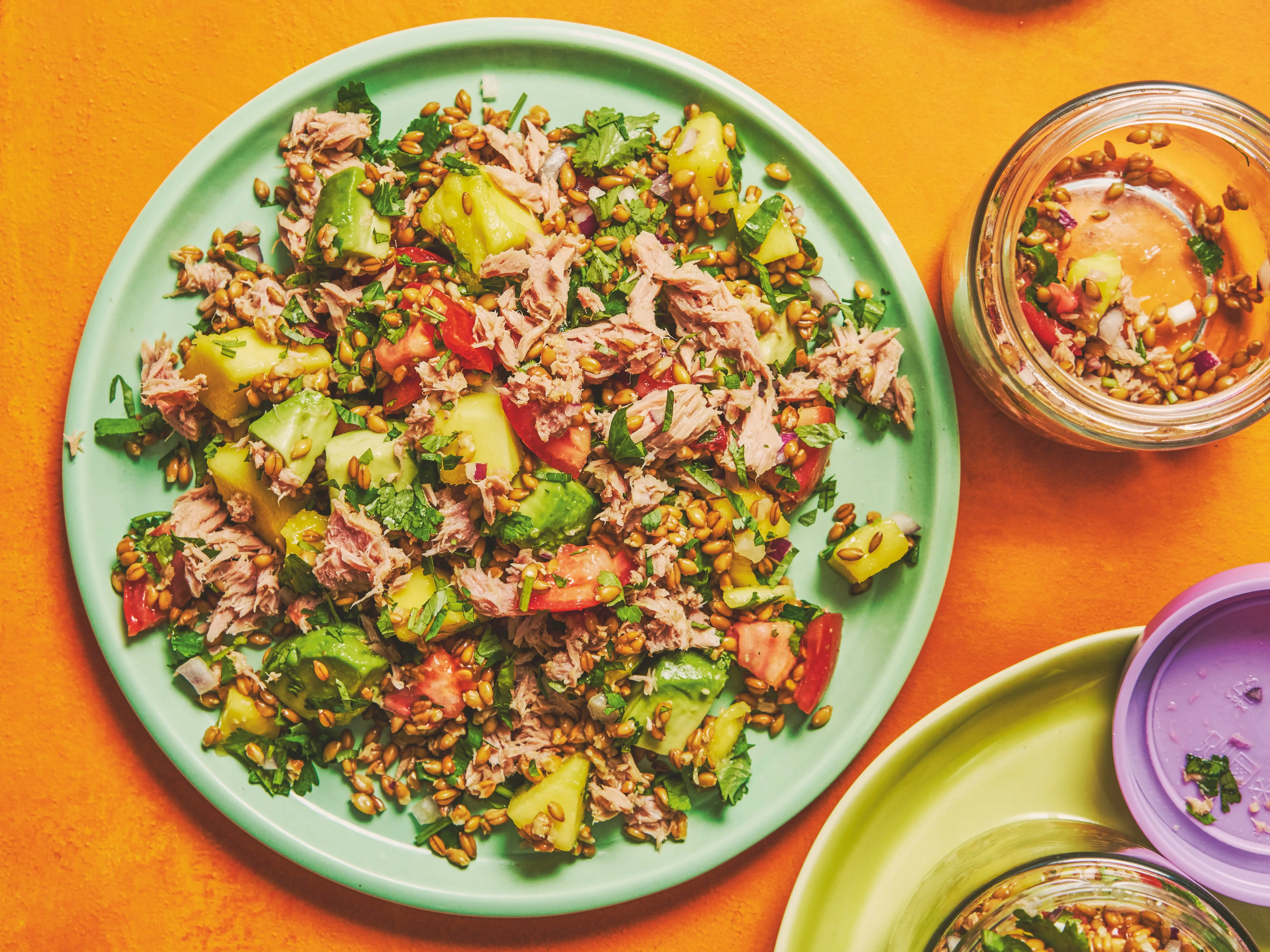Overhead view of a green plate with shredded fish, grains, cucumber, tomatoes, herbs, and jars on an orange surface.