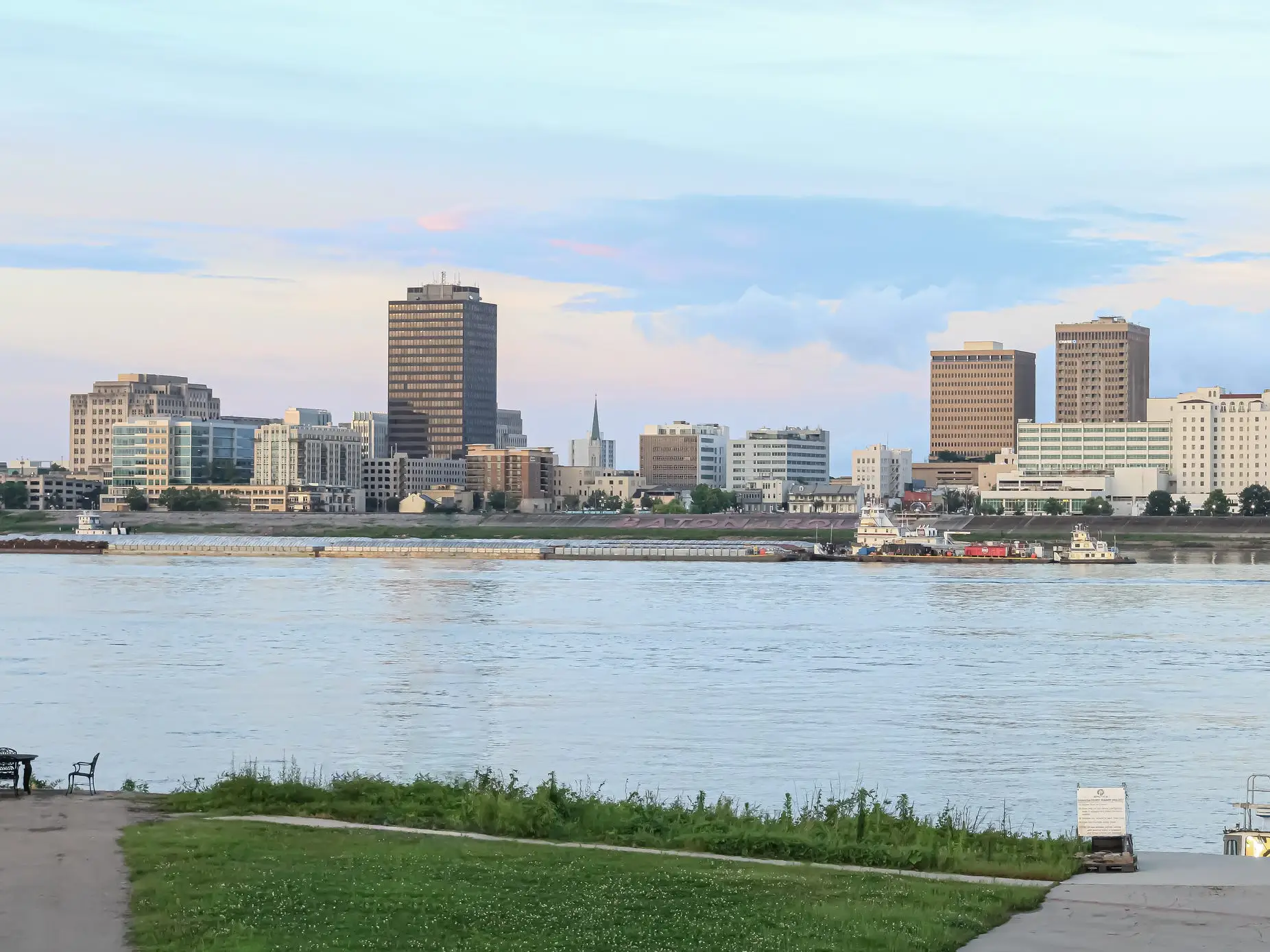 Barges pass the Baton Rouge skyline across the Mississippi River beneath a pastel evening sky.