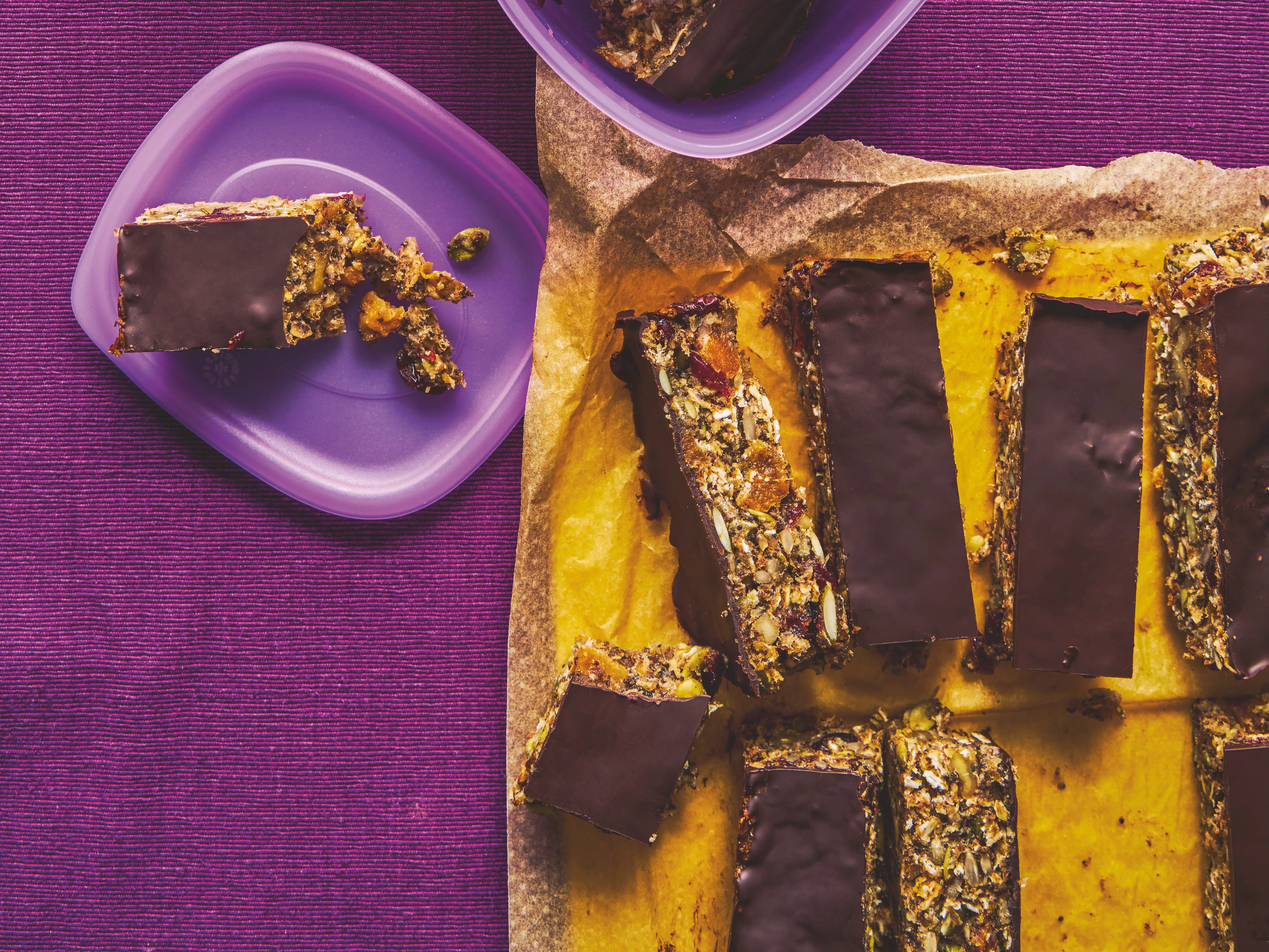 Chocolate-topped snack bars sit in plastic containers and on parchment paper over a purple textured cloth.