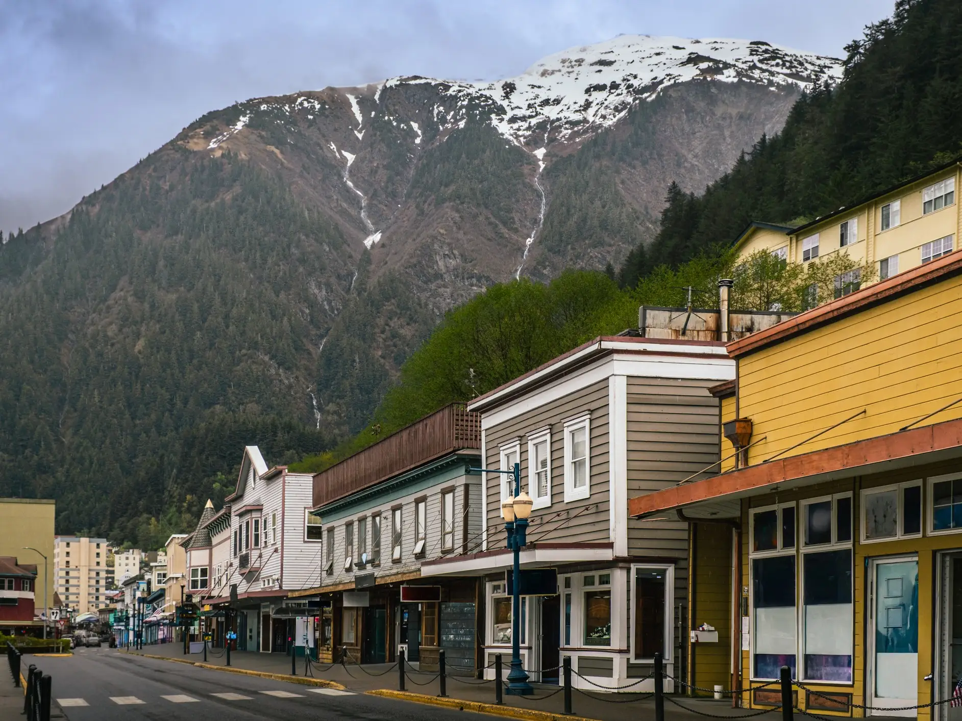 Colorful storefronts line a quiet street below a forested mountain with patchy snow under cloudy skies.