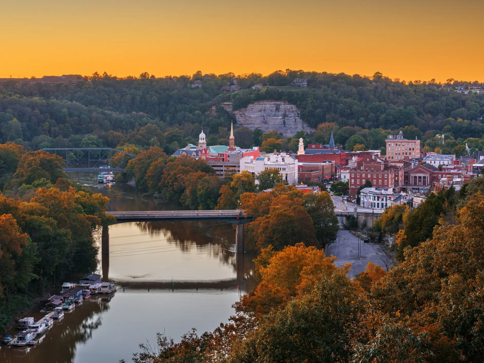 Downtown Frankfort and the Kentucky River sit among autumn trees under an orange sunset sky.