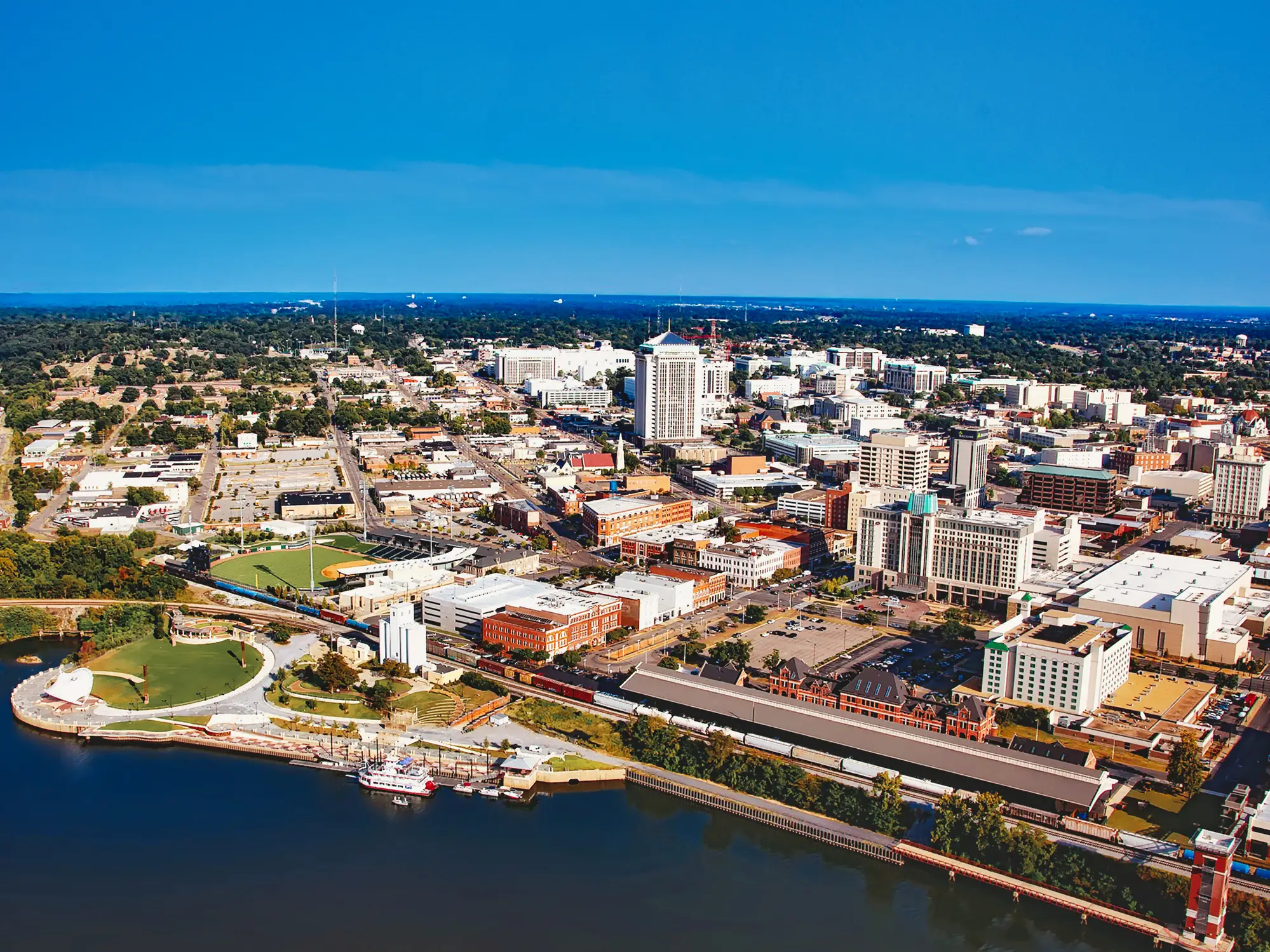 Aerial view of downtown Montgomery beside the Alabama River, with skyline, rail tracks, riverfront park, and baseball stadium.