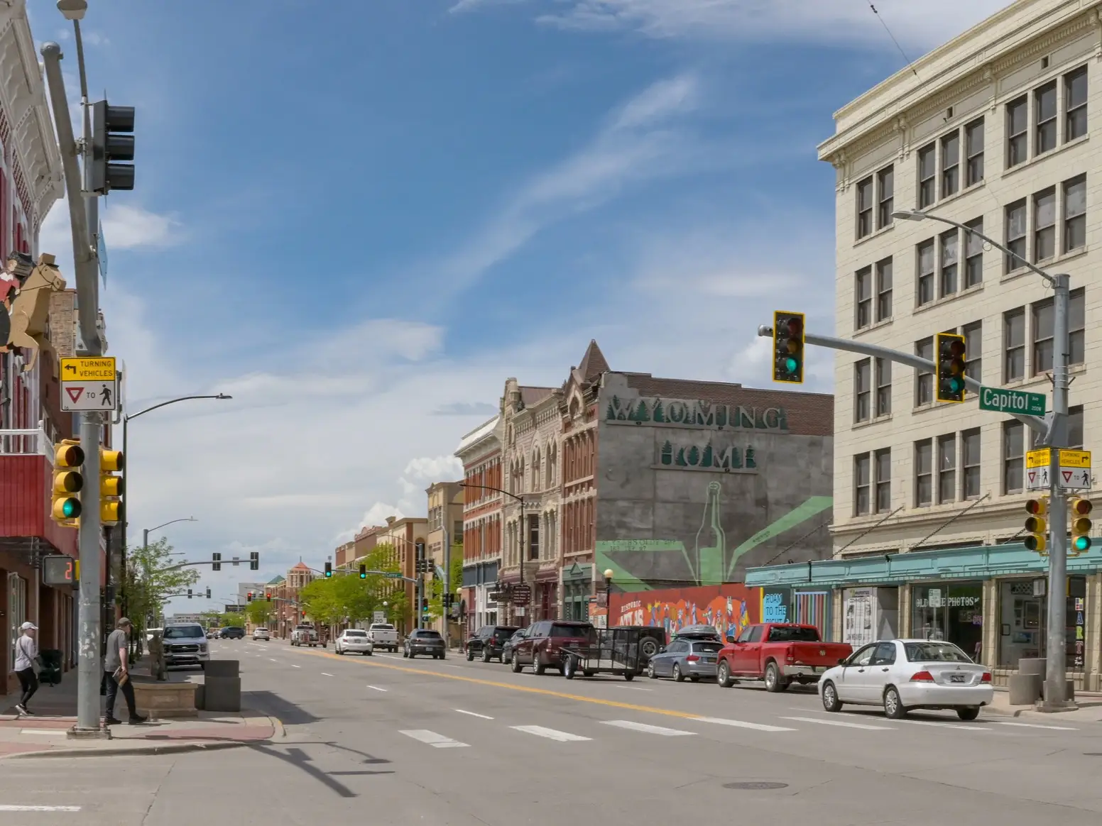 Downtown street intersection with traffic lights, parked cars, storefronts, and a large “WYOMING HOME” mural.
