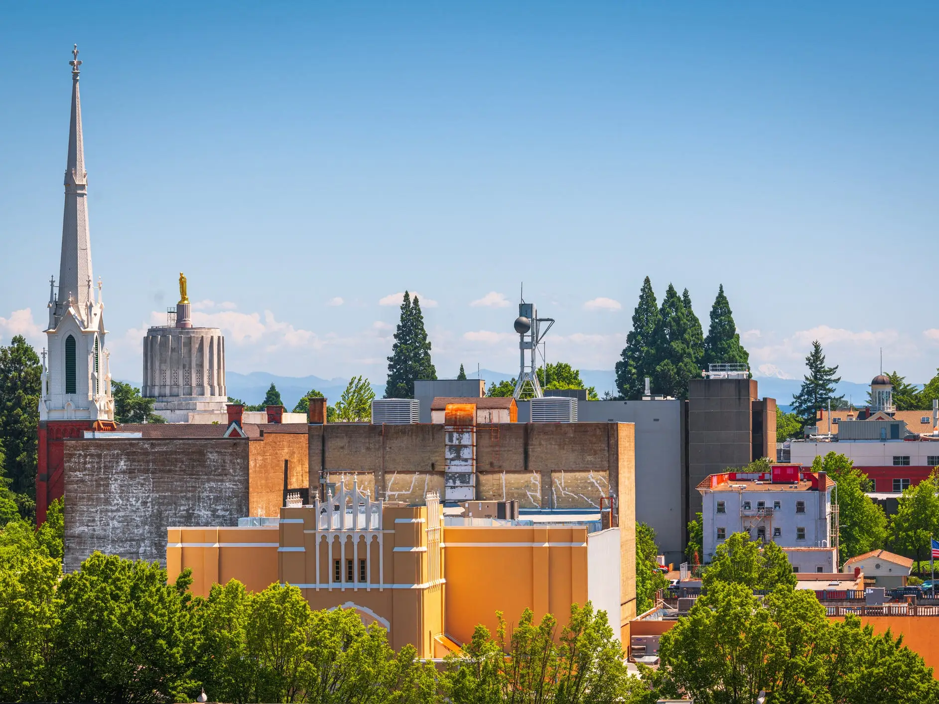 Downtown Salem skyline with a church steeple, trees, and the Oregon State Capitol dome under a clear blue sky.