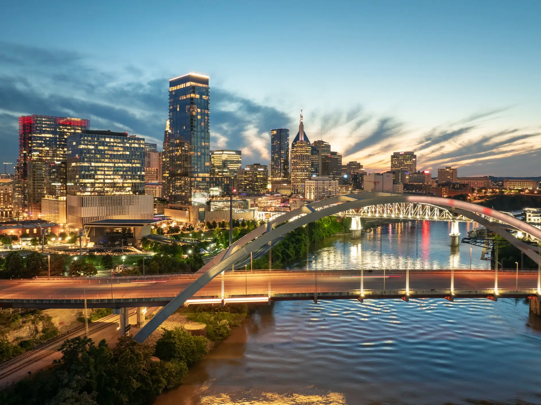 Downtown Nashville skyline glows at dusk behind an arched bridge over the Cumberland River.
